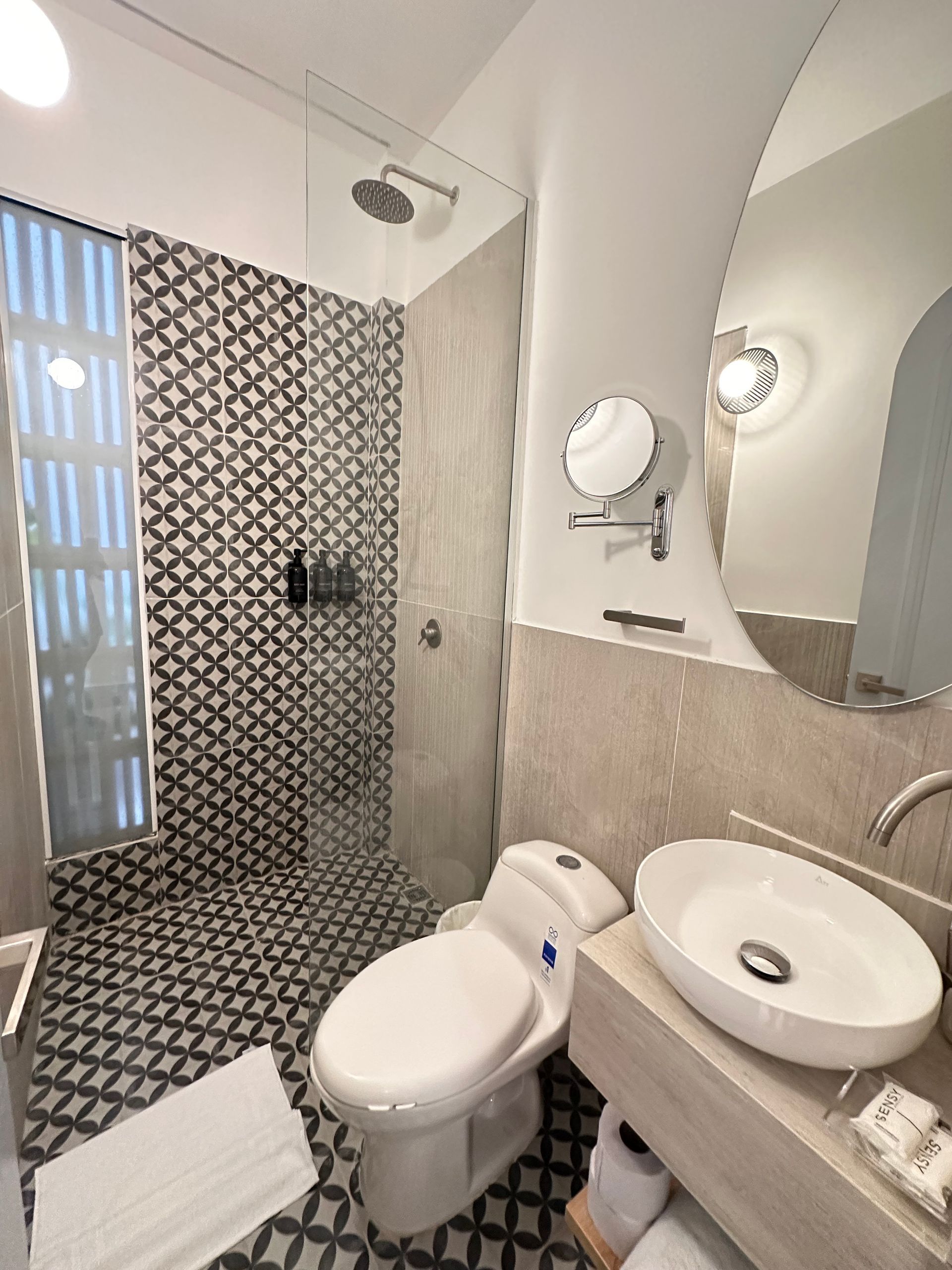 Modern bathroom with black and white patterned tile, a shower, toilet, and a sink with a large, oval mirror.
