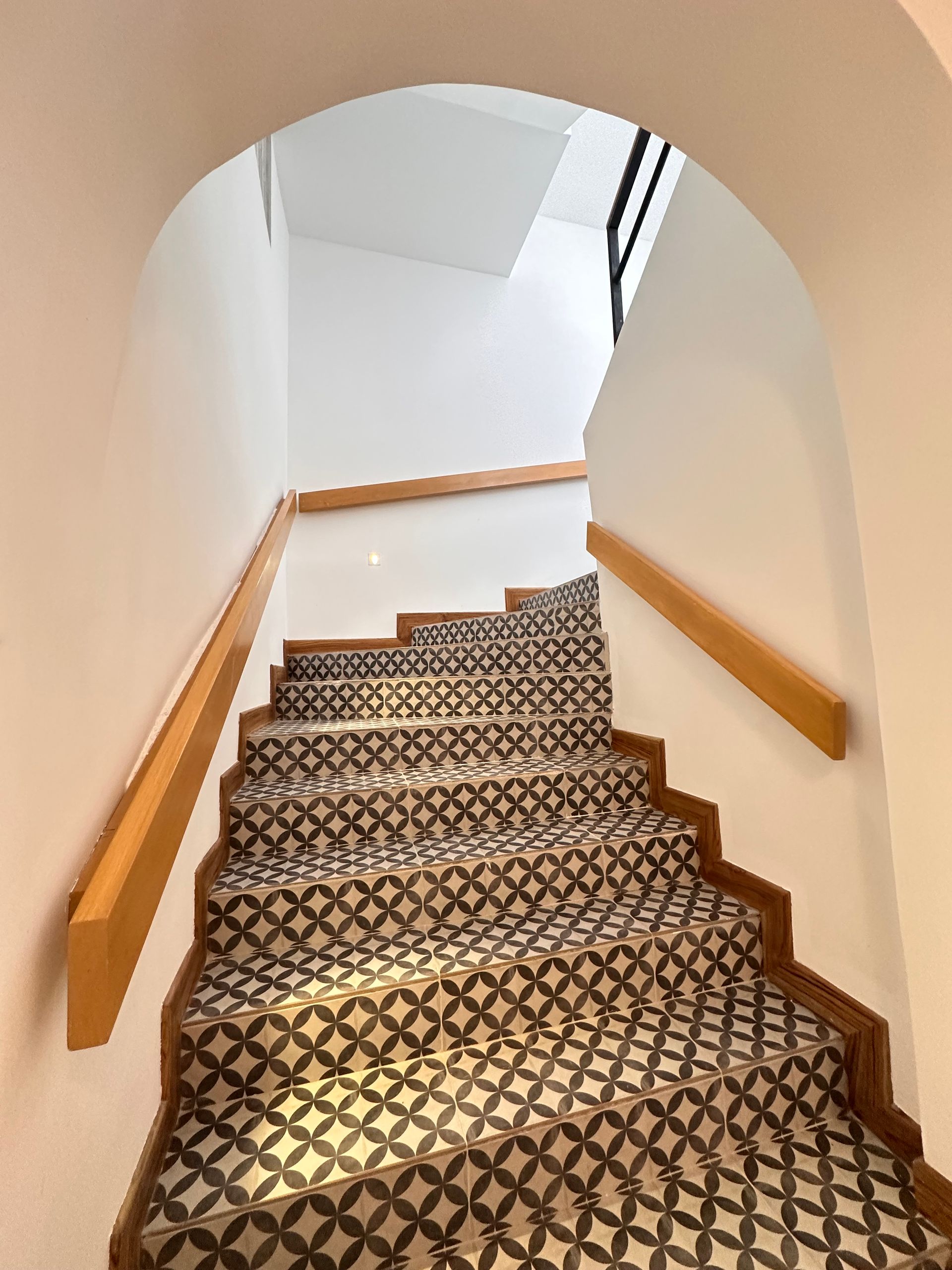 Staircase with patterned tile steps ascends under an arched opening. Wooden handrails line both sides, walls are white.