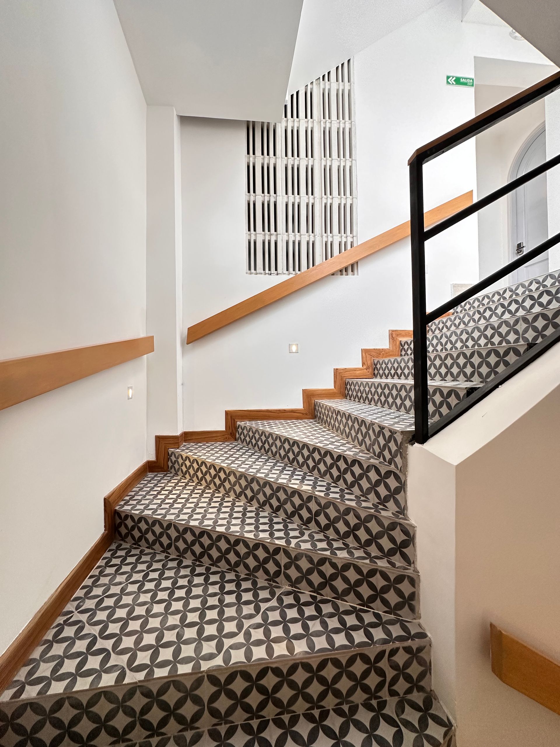 Staircase with patterned tiles and wooden handrails, leading upwards. Features a black metal railing and decorative window.