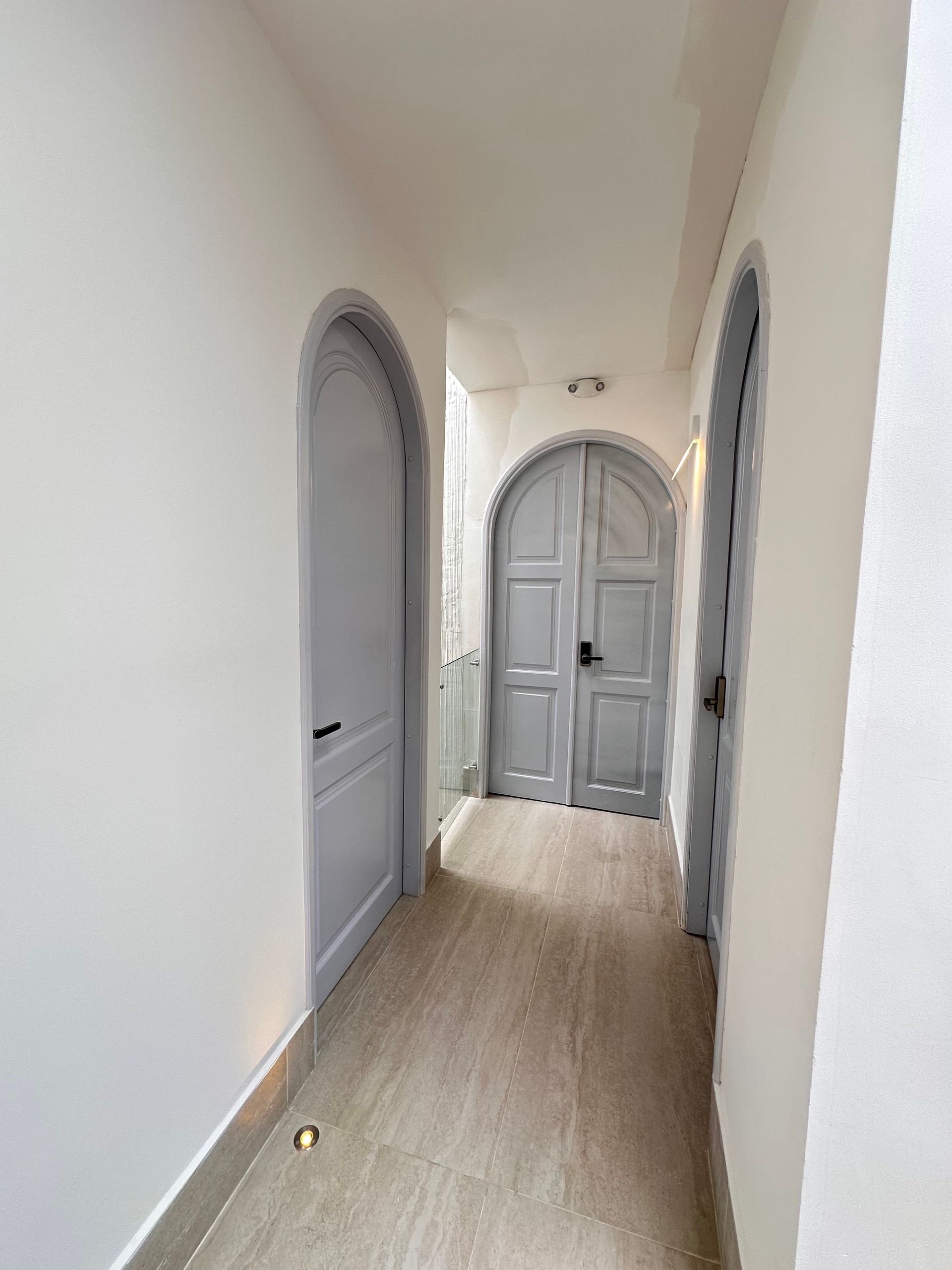 Narrow hallway with white walls and arched, gray doors. The floor is light-colored with in-ground lights.