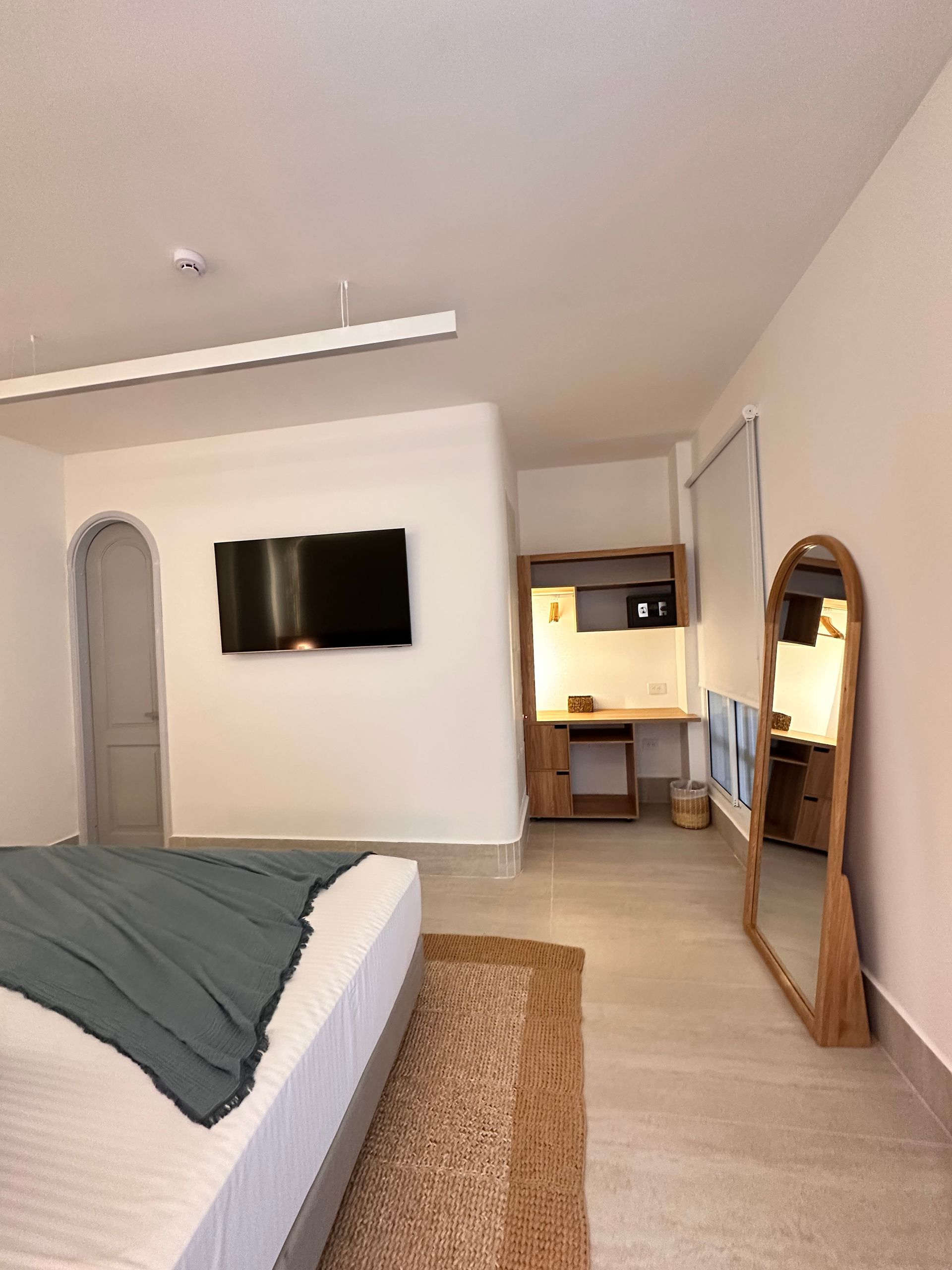 A modern, bright bedroom with a bed, TV, desk, and full-length mirror. The room is white with natural wood accents.