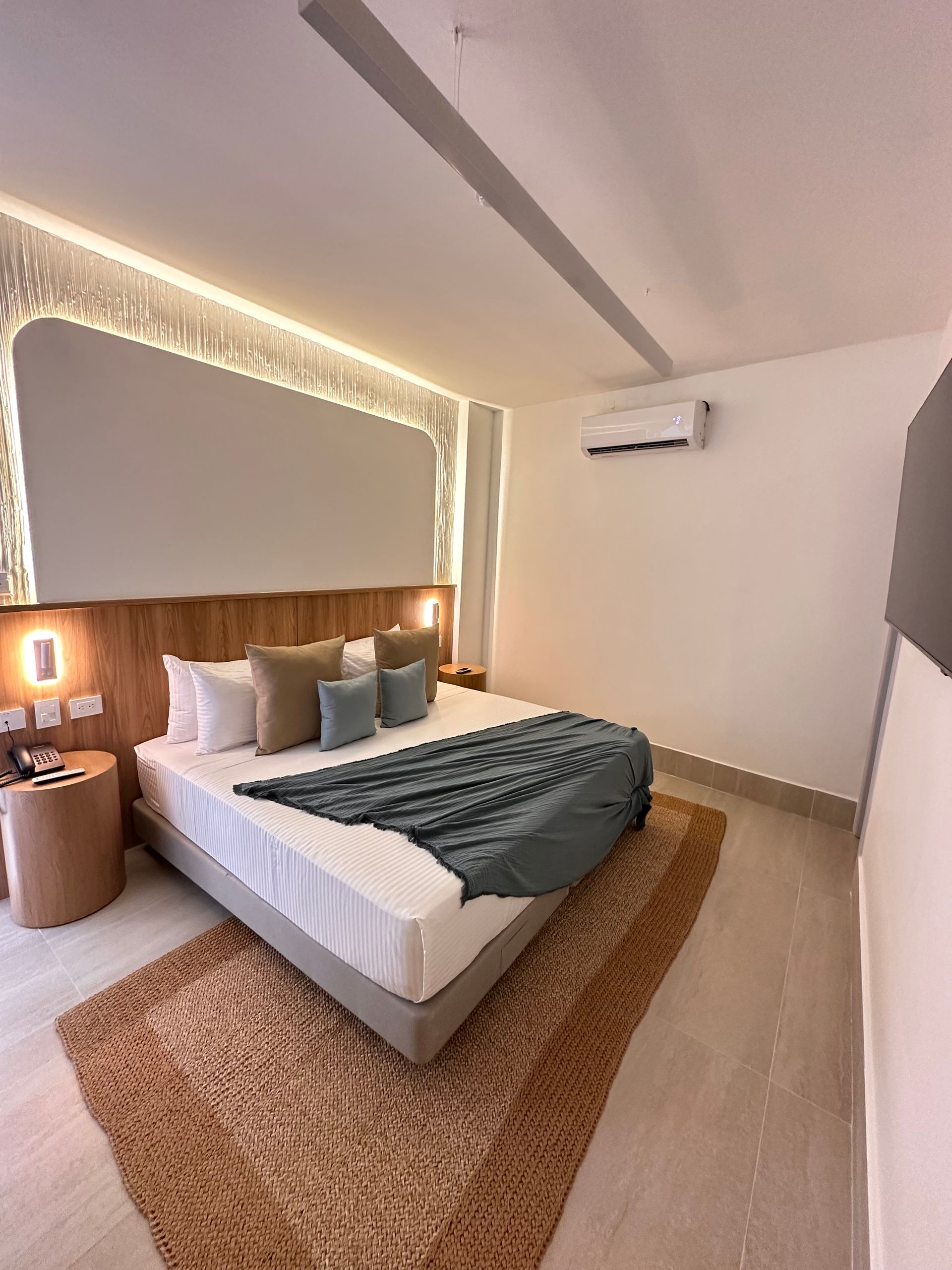 A hotel bedroom with a bed covered in pillows and a teal blanket, a wooden headboard, and a decorative rug.