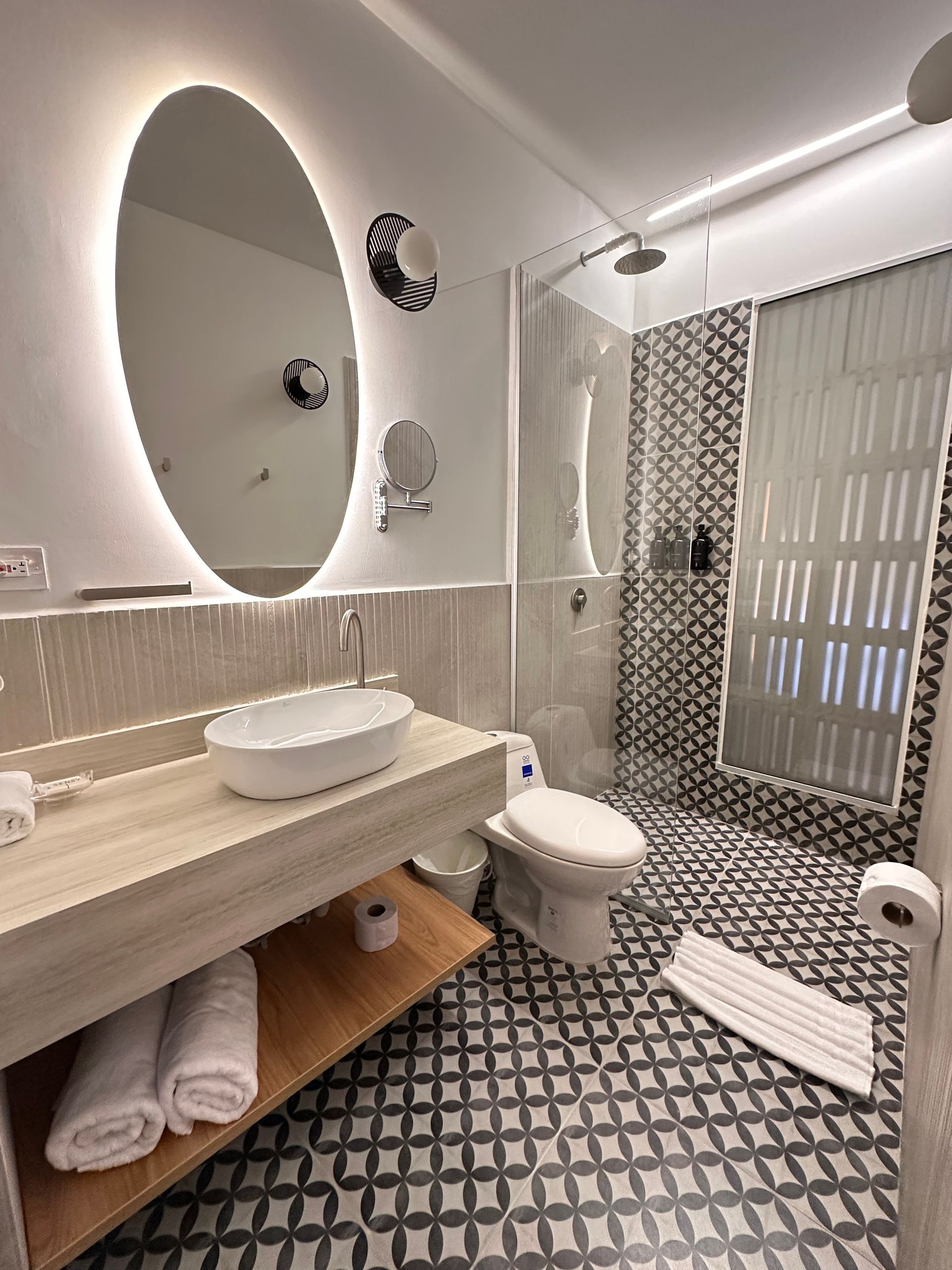Modern bathroom with a white sink, toilet, and shower. Black and white patterned floor and wall tiles, large oval mirror with light.