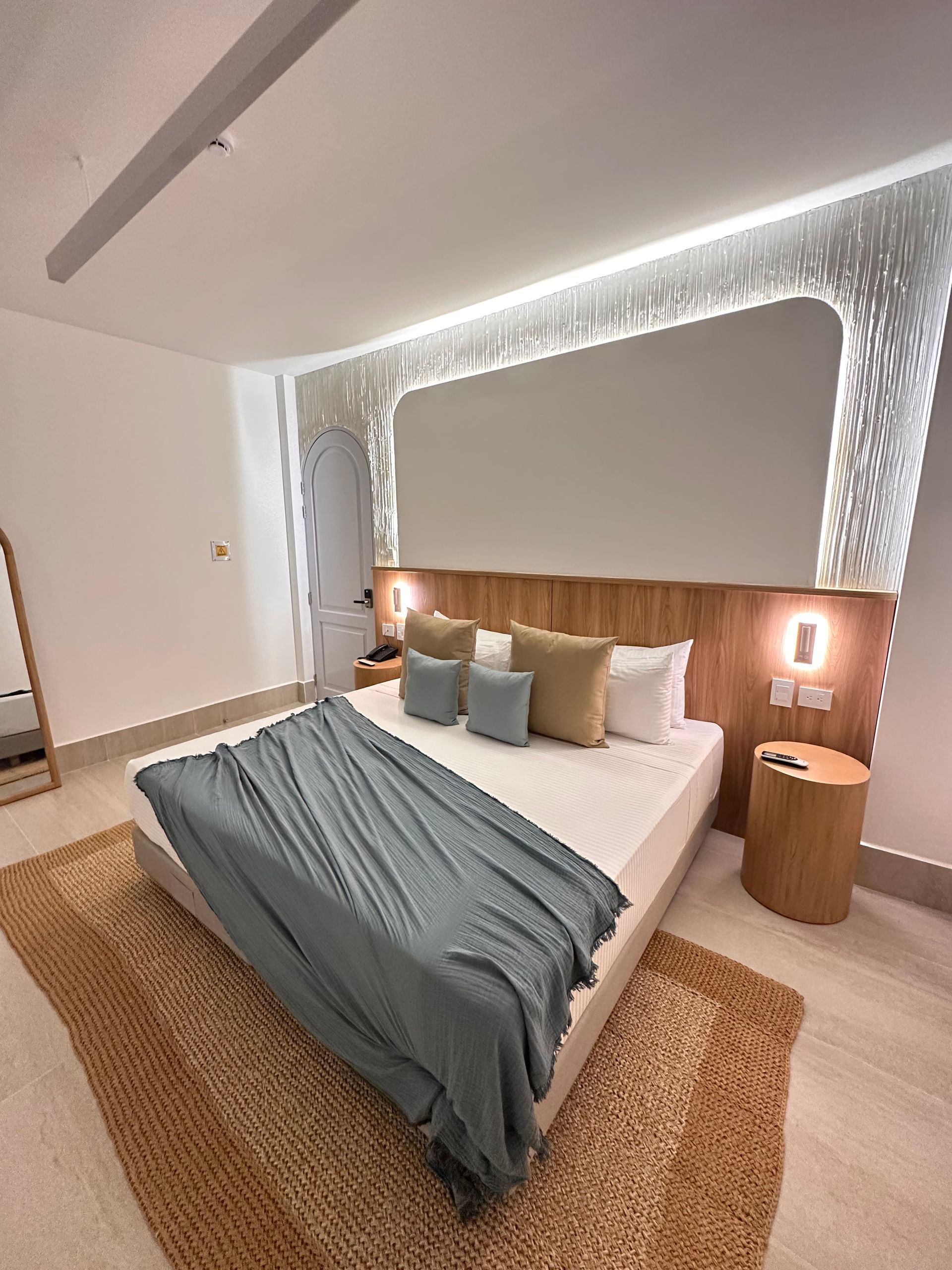 Cozy hotel bedroom with a large bed, wooden headboard, textured wall, and woven rug. Soft blue and beige accents create a relaxing ambiance.