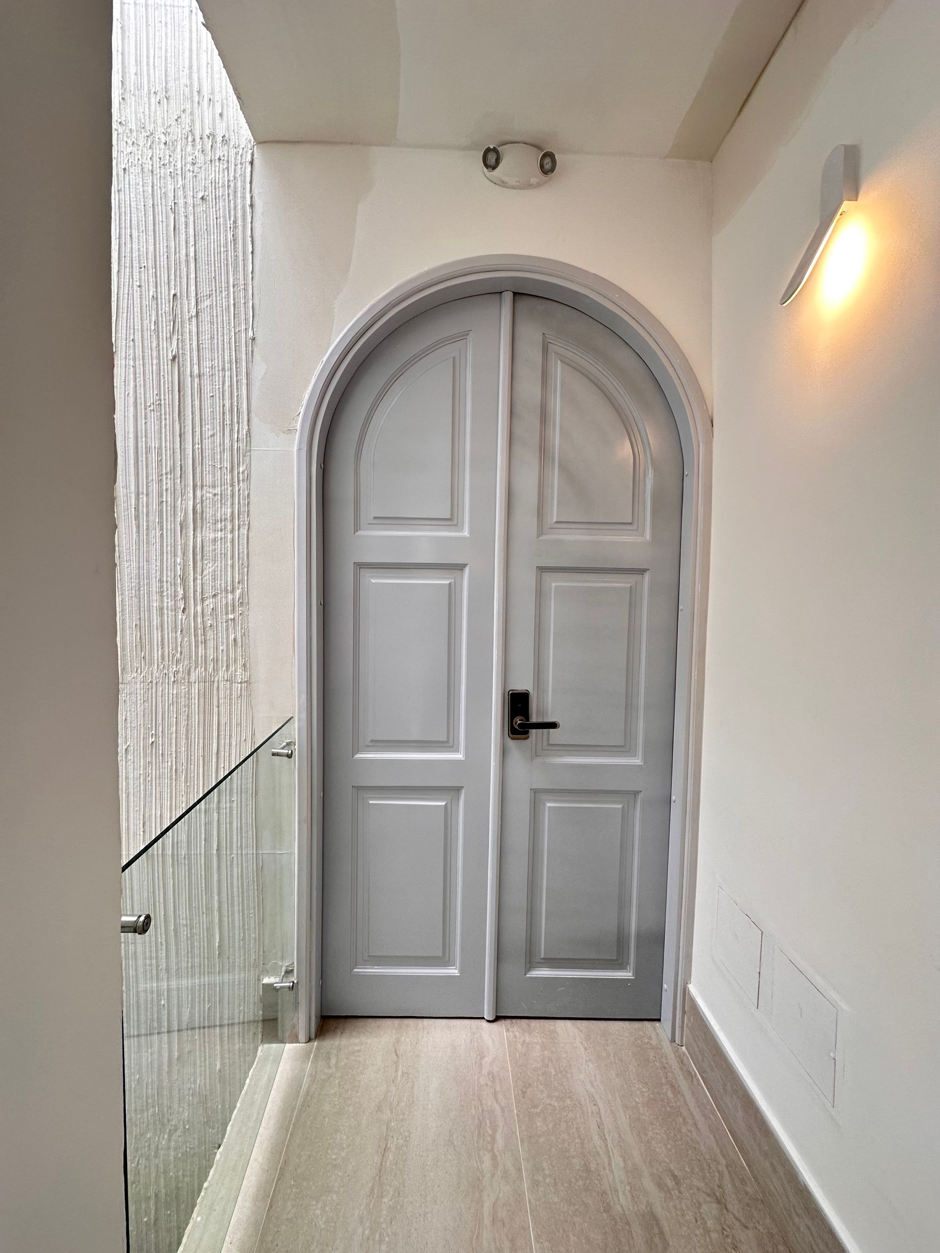 Gray arched double doors in a white hallway with a wood-look floor. A small light is above the doors, with a side light to the right.