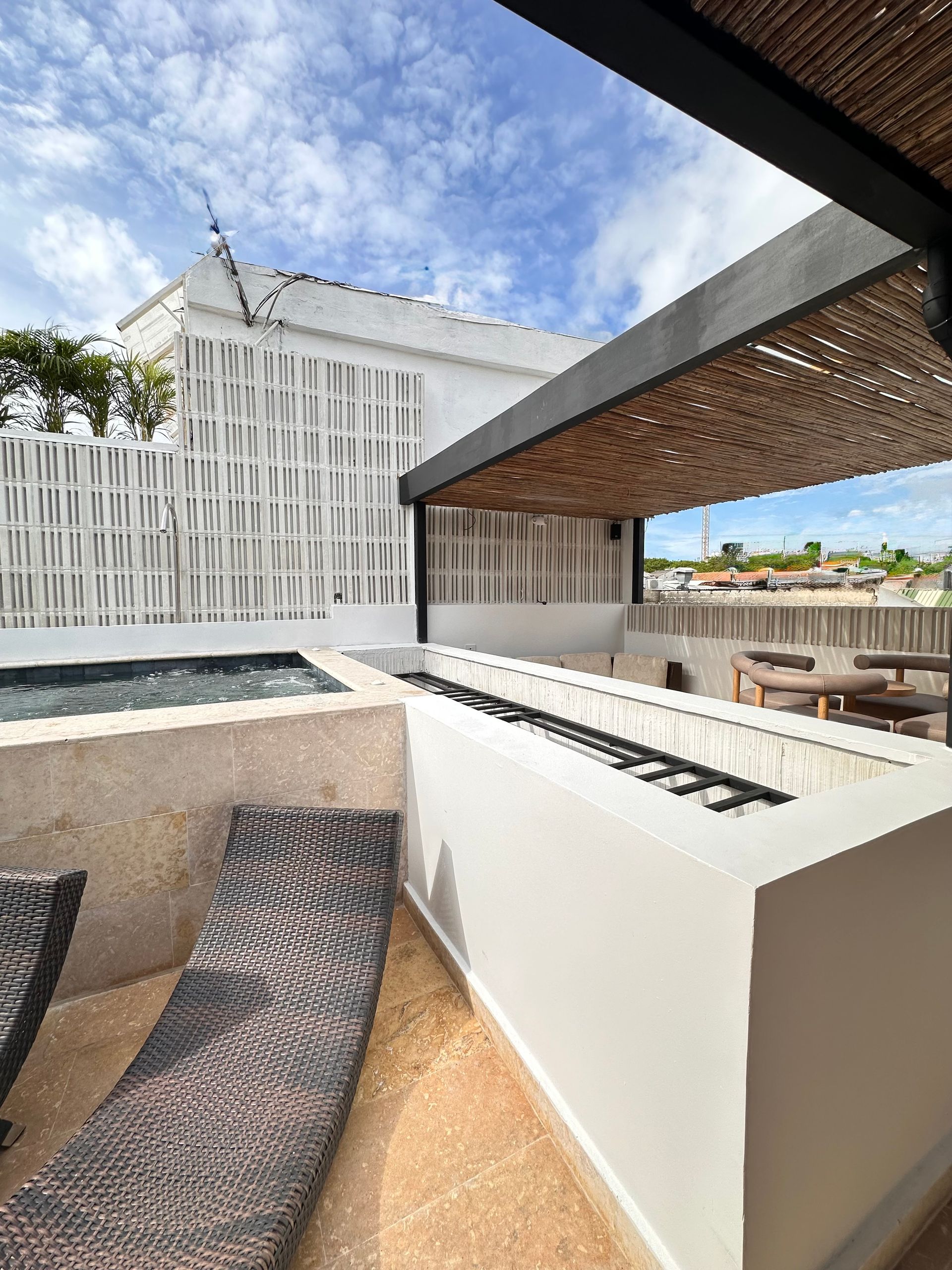 Rooftop terrace with a pool, lounge chairs, and a woven-roof pergola; white buildings and a blue sky are visible.