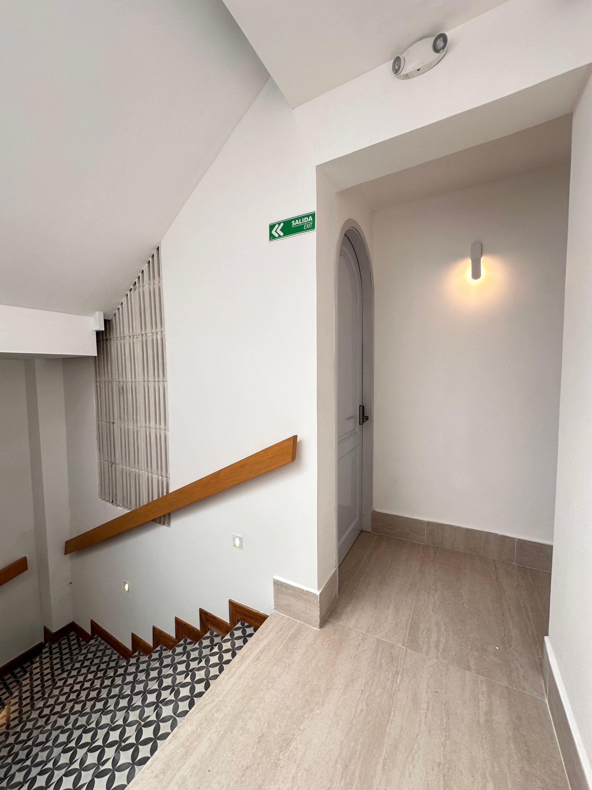 Stairwell with patterned steps, a wooden handrail, and a door at the end of a hallway. White walls, a green exit sign, and a lit wall sconce.