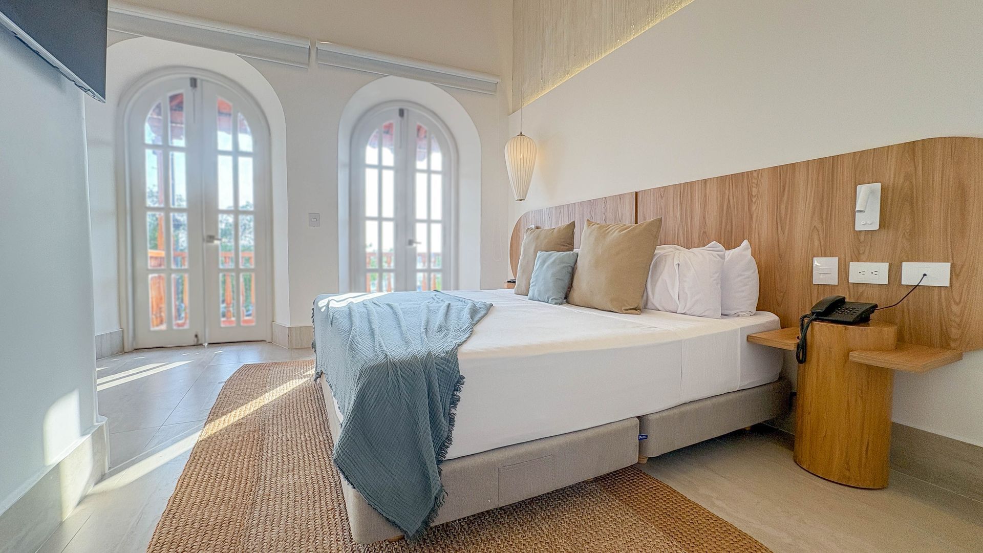 Bright hotel bedroom with a large bed, light wood headboard, and two arched French doors leading to a balcony.