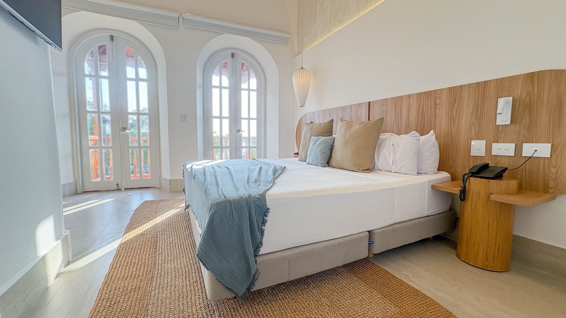 Bright bedroom with white walls, arch windows, and a bed with pillows and a blue blanket. Wooden headboard and side table.