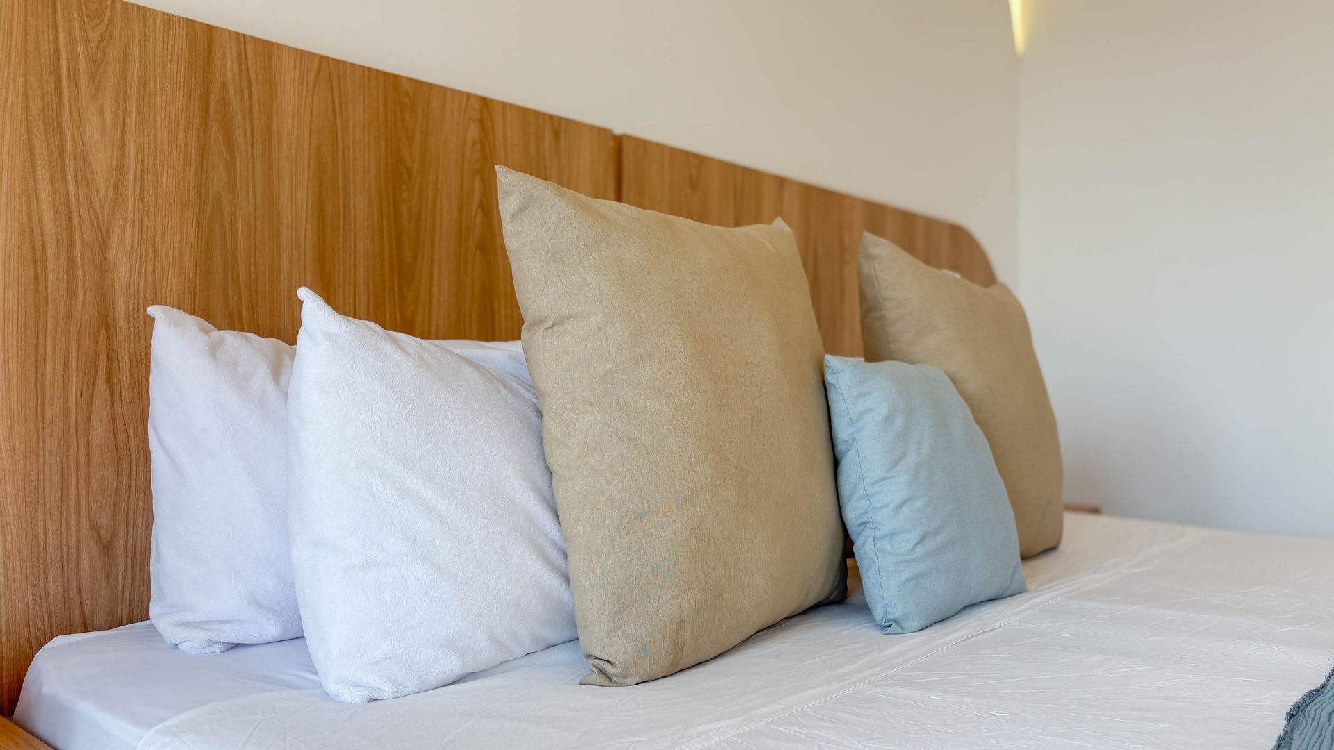 Bed with white bedding and pillows in various colors against a wooden headboard and white wall.
