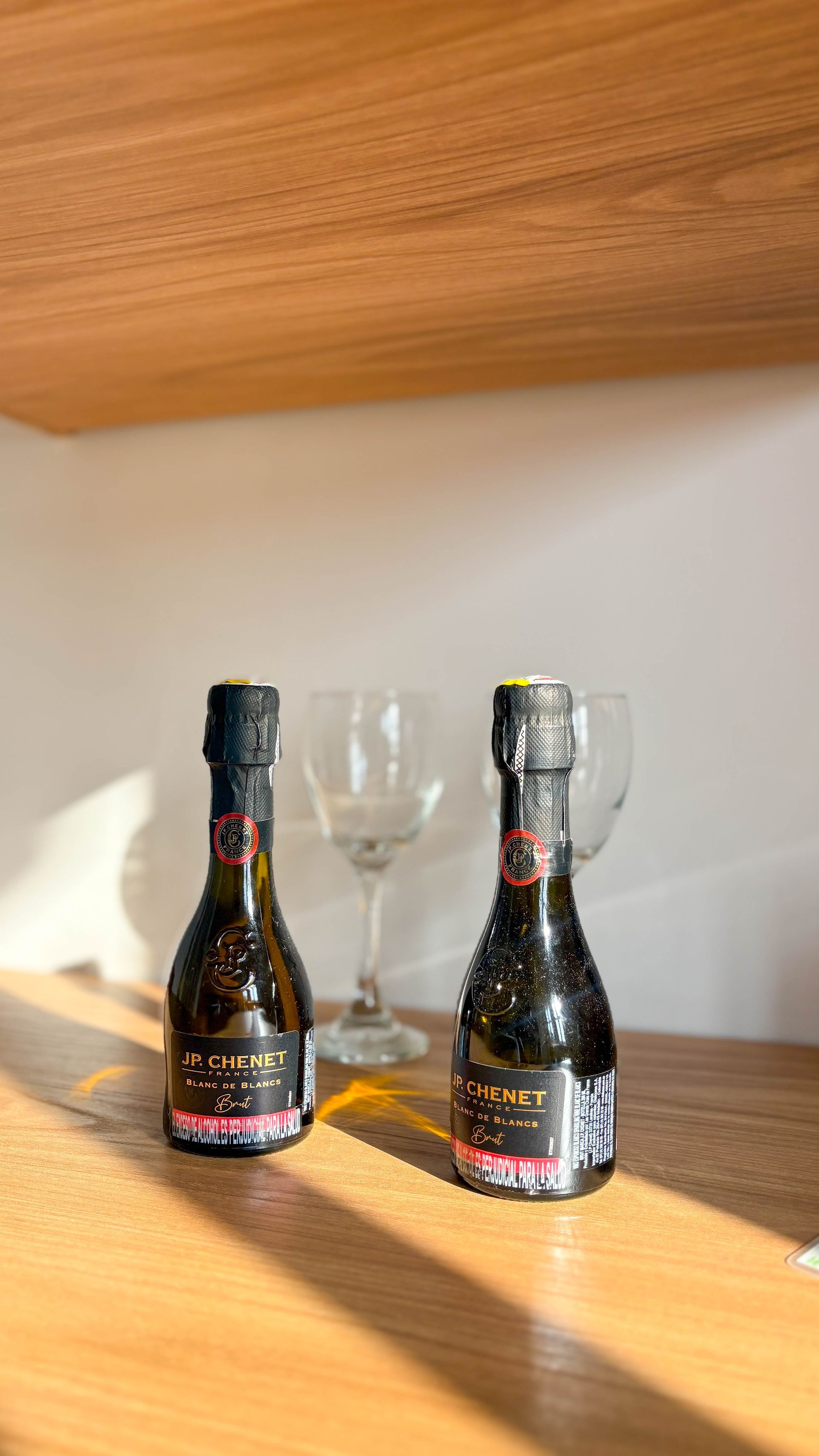 Two small, dark bottles of sparkling wine stand on a wooden shelf, with a glass behind them. Sunlight casts shadows.