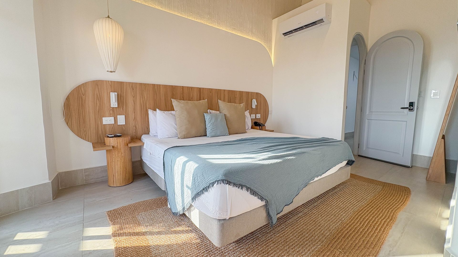 Bedroom with a large bed, wooden headboard, and natural-toned rug. Soft blue blanket and pillows. Bright, airy interior.