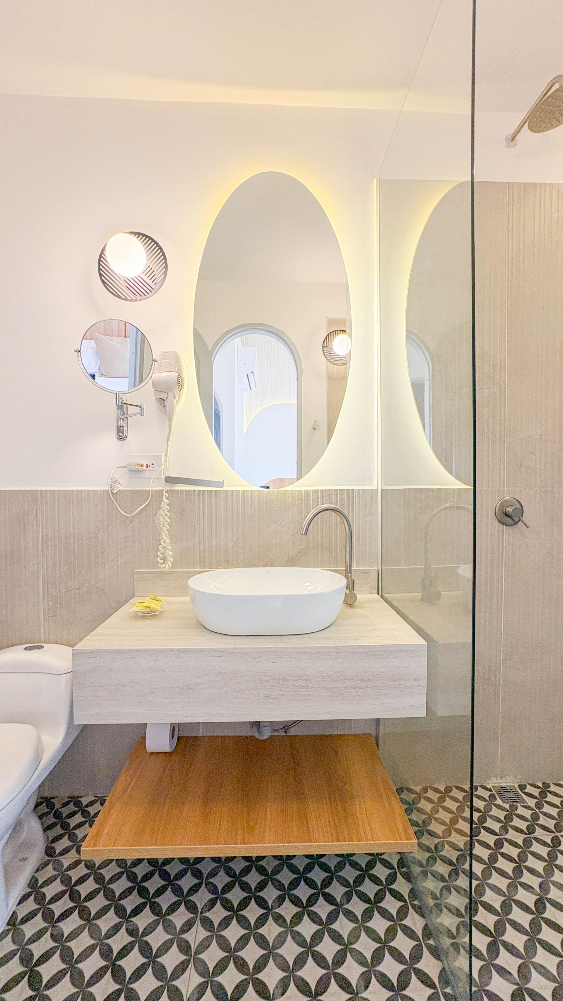 Modern bathroom with a floating sink, large oval mirror with a backlit frame, and patterned tile flooring.