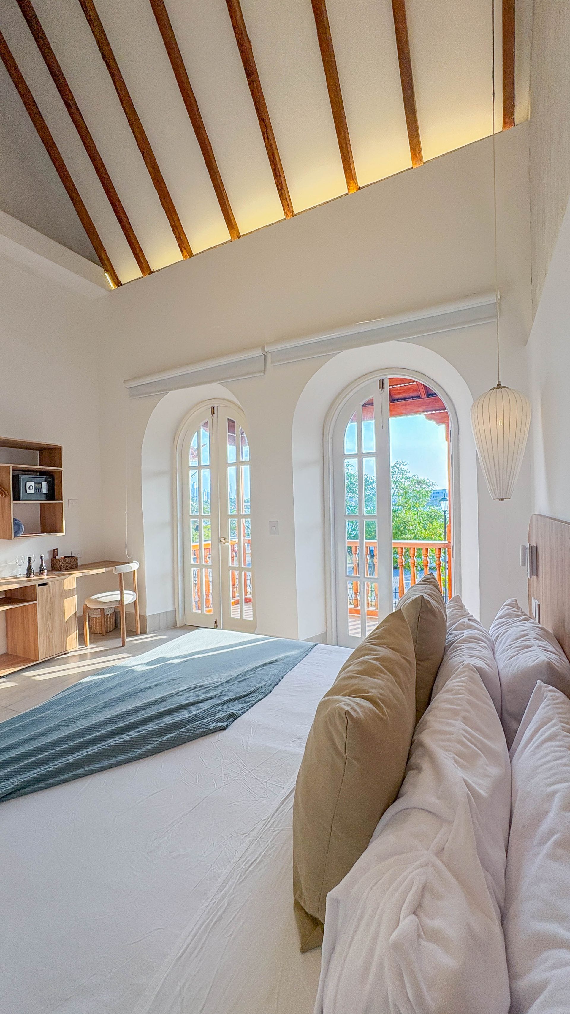 Bedroom with white walls and bed, archway windows lead to a balcony with a view of trees and blue sky.