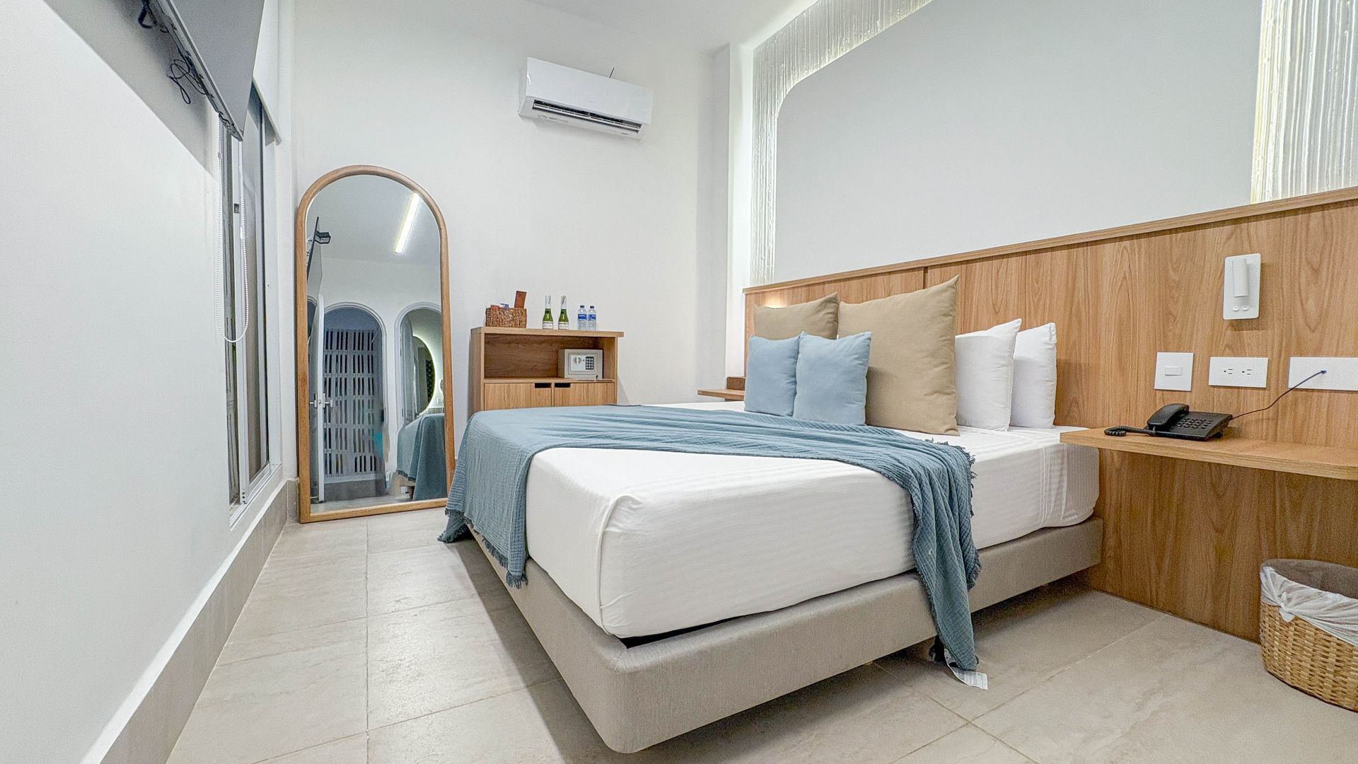 A modern hotel room with a queen-sized bed, large mirror, and white walls. Blue accents and wood furniture create a relaxing atmosphere.