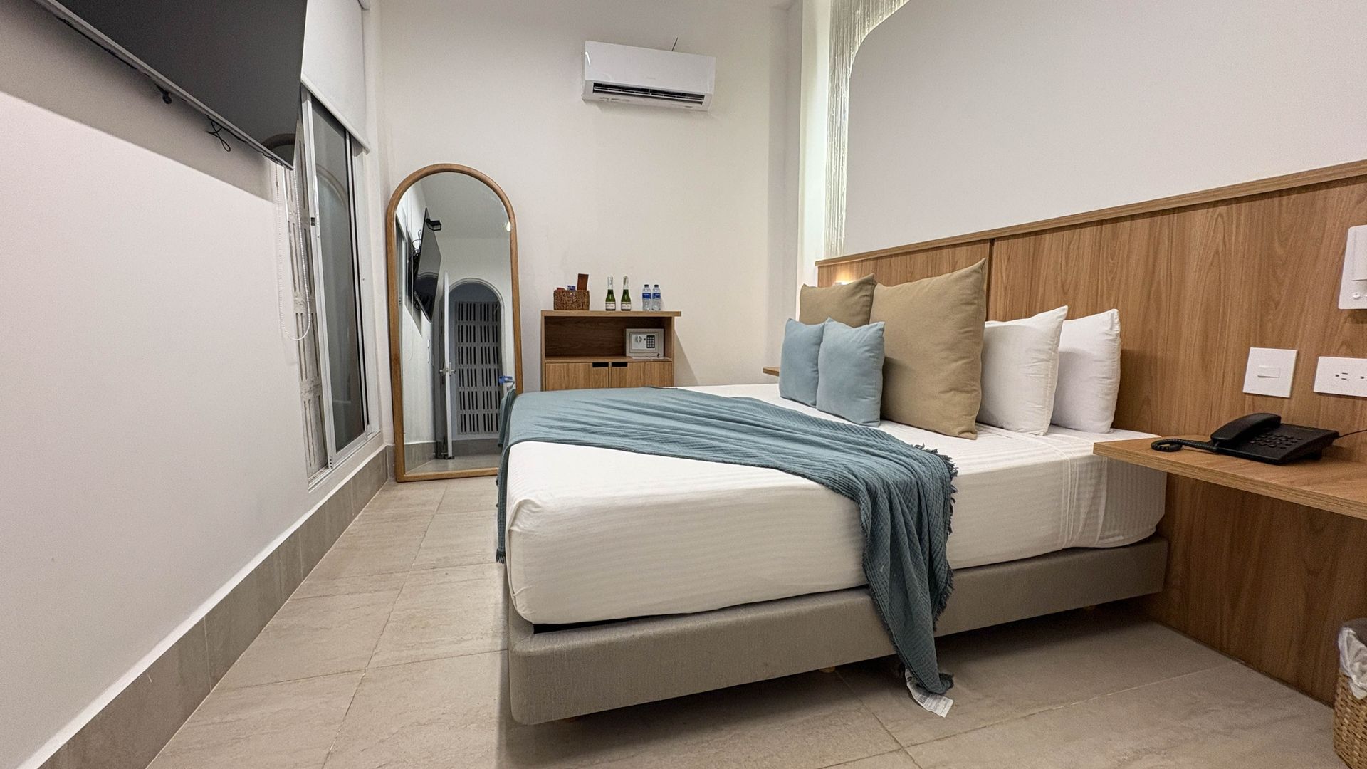 A modern hotel room with a bed, large mirror, and small desk. The room is white and wood-toned with light blue accents.