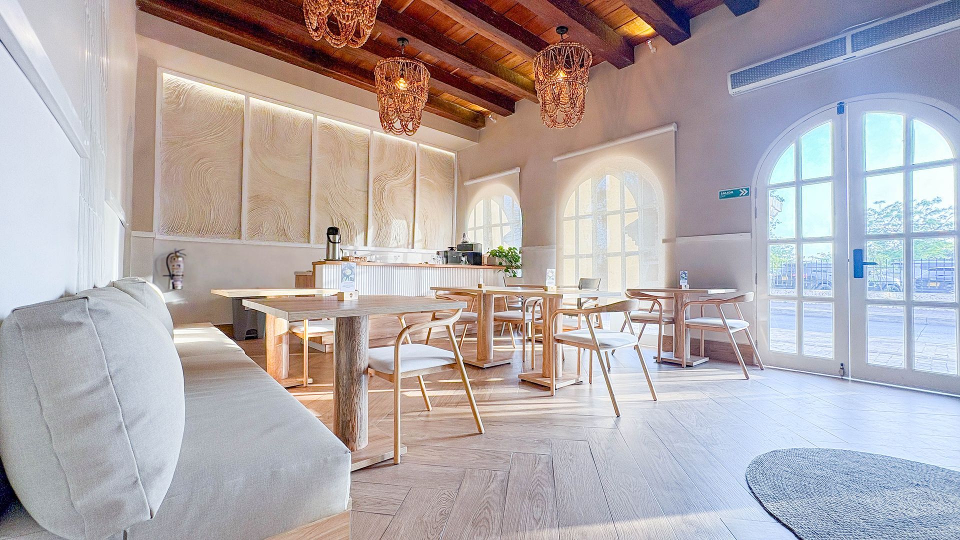 Bright, airy dining room with light wood furniture, arching windows, and decorative hanging lights. White walls and a large white sofa are also present.