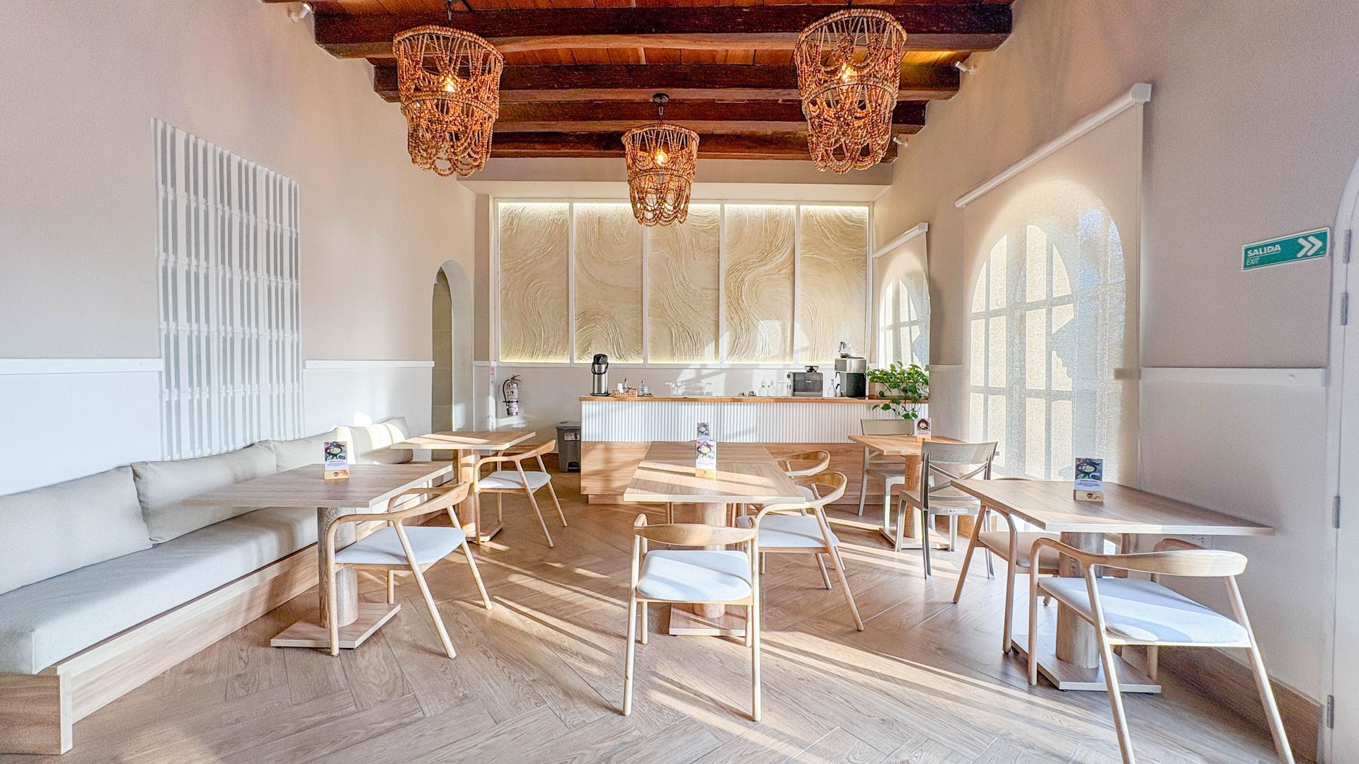 Bright, airy restaurant interior with wooden tables, chairs, and decorative lighting. White walls and a long counter with a patterned backdrop.