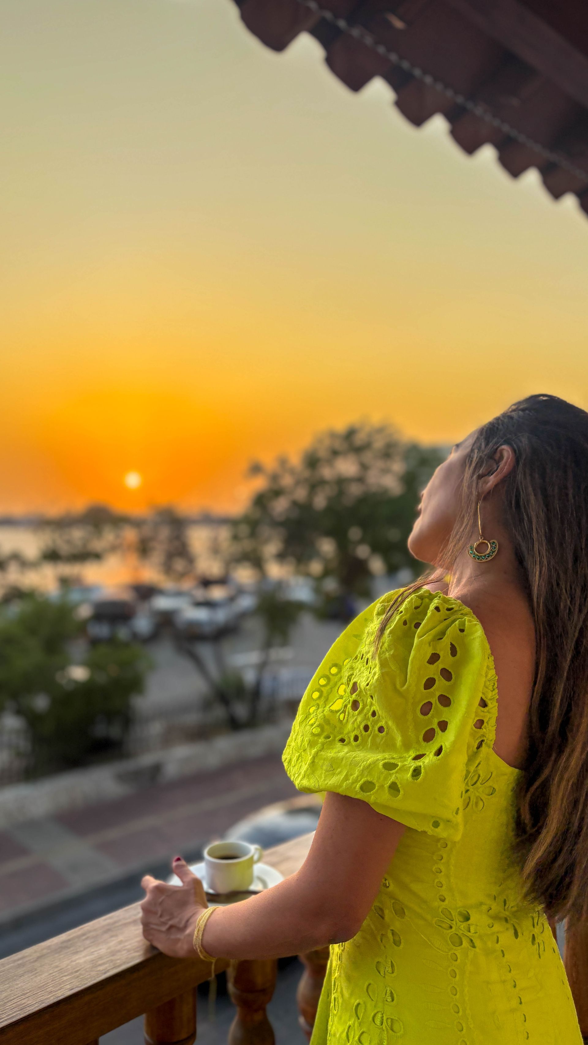 Woman on a balcony looks at the sunset over water. She wears a yellow dress and holds a cup.