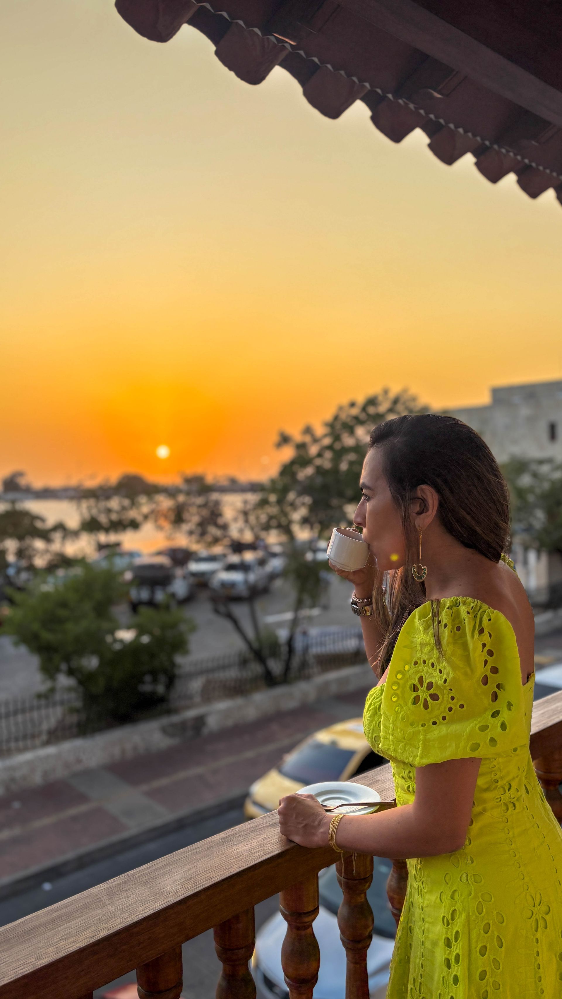 Woman in neon yellow dress sips from a cup, looking out at sunset over water. Balcony setting, gold accessories.