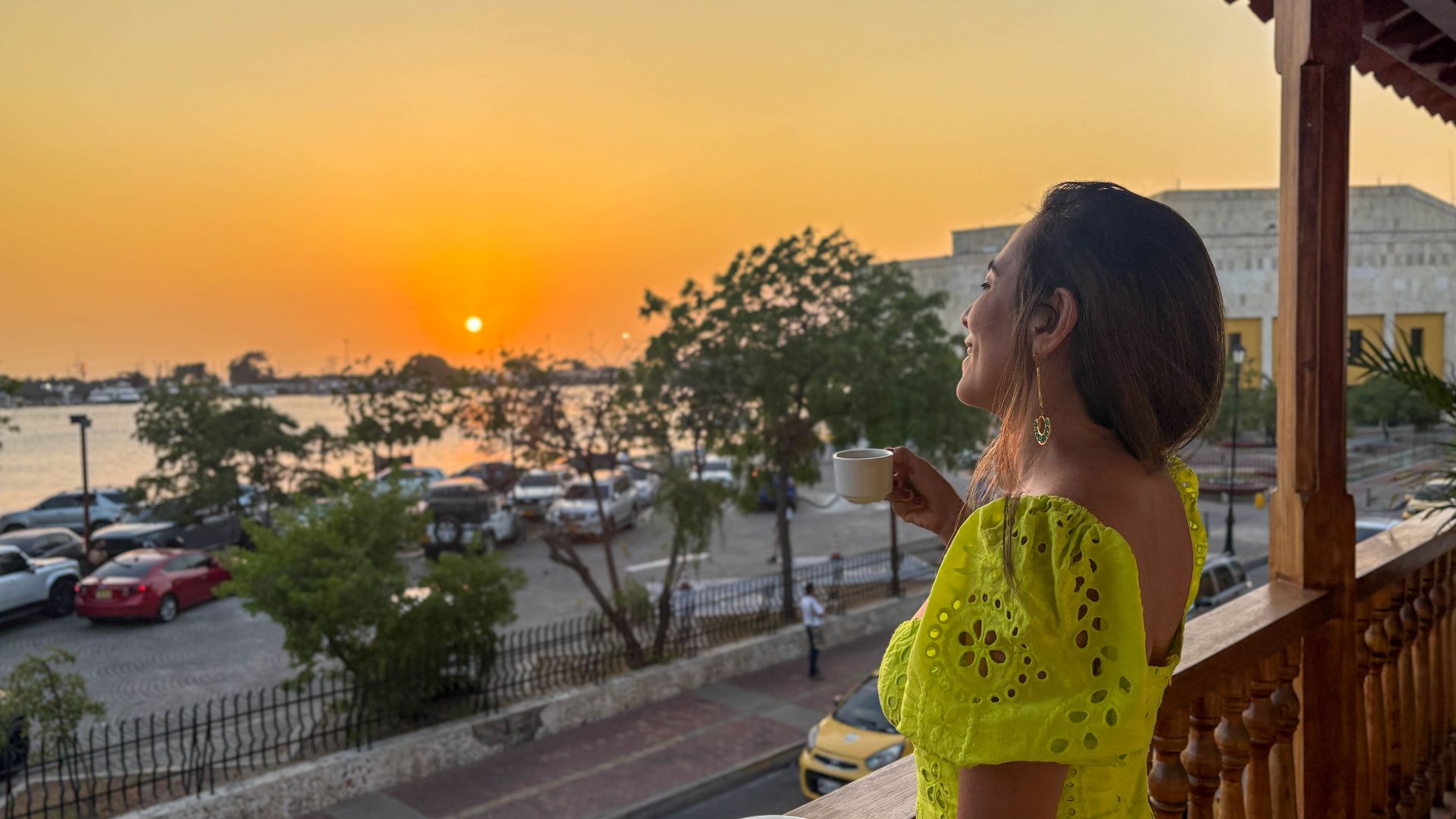 Woman on balcony enjoying sunset with a cup of coffee. Overlooking water and parked cars. Wearing neon yellow dress.