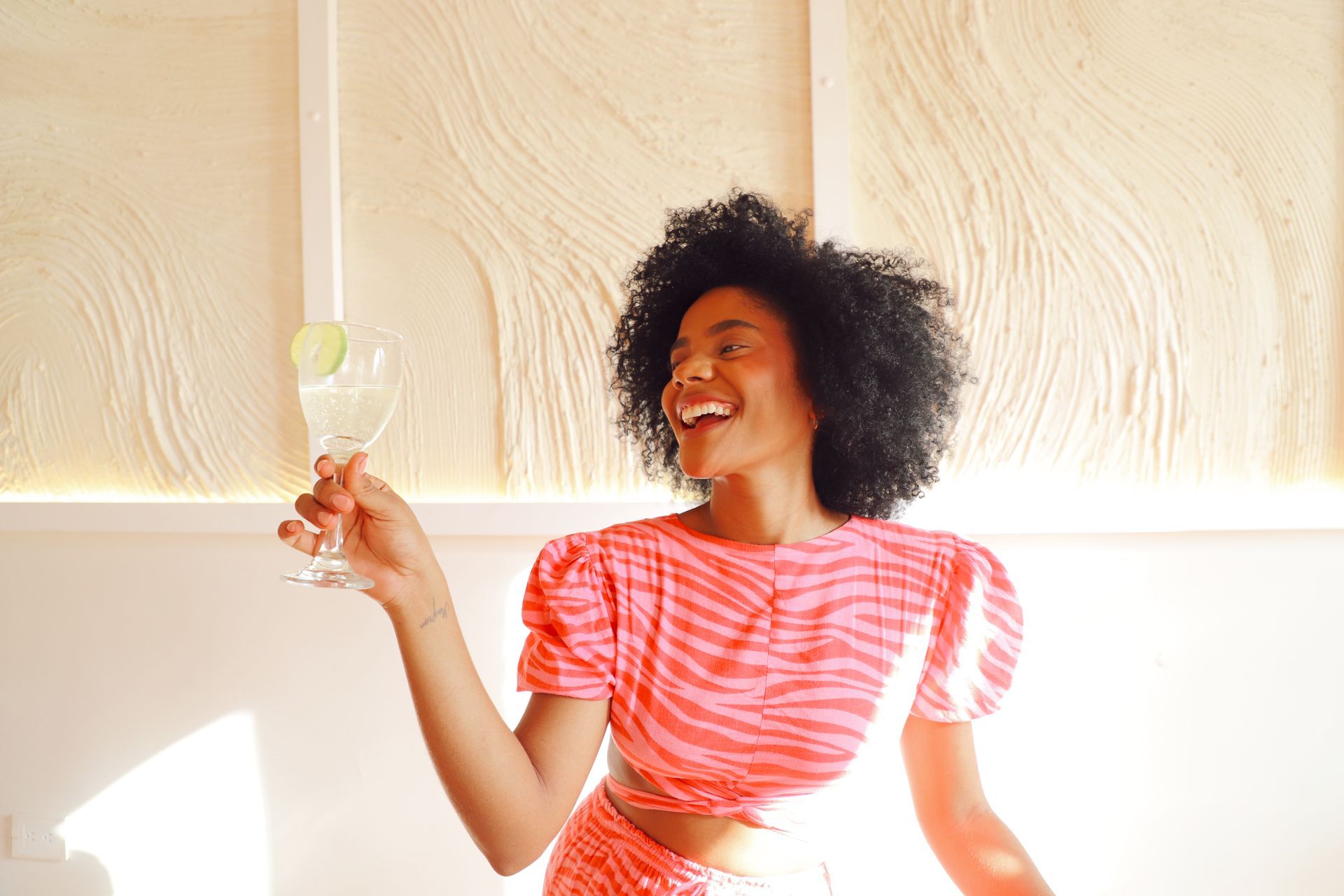 Woman with dark curly hair, wearing a pink zebra-print outfit, laughing and holding a cocktail with a lime slice, indoors.