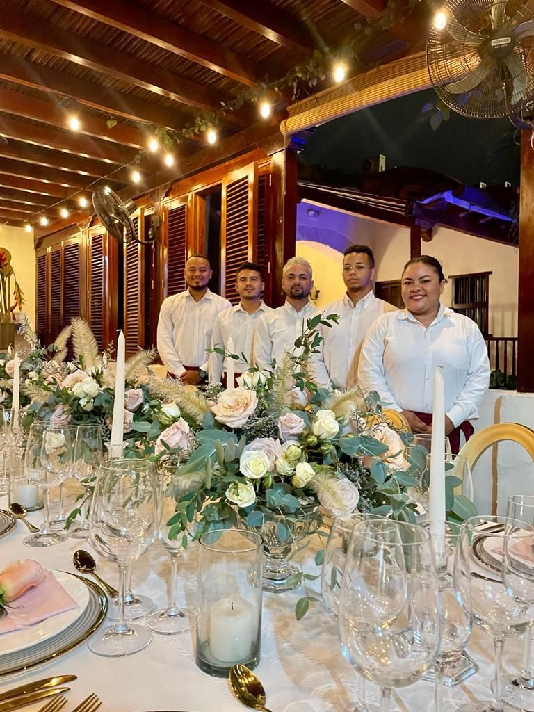 Five servers in white shirts stand behind a beautifully decorated table set for a special occasion; flowers and candles adorn the table.