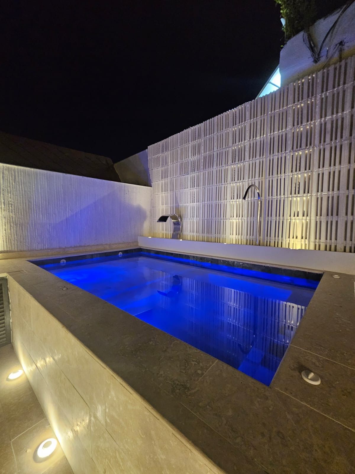 A rectangular pool at night, illuminated blue. Beige stone surrounds the pool with a white, latticed wall in the background.