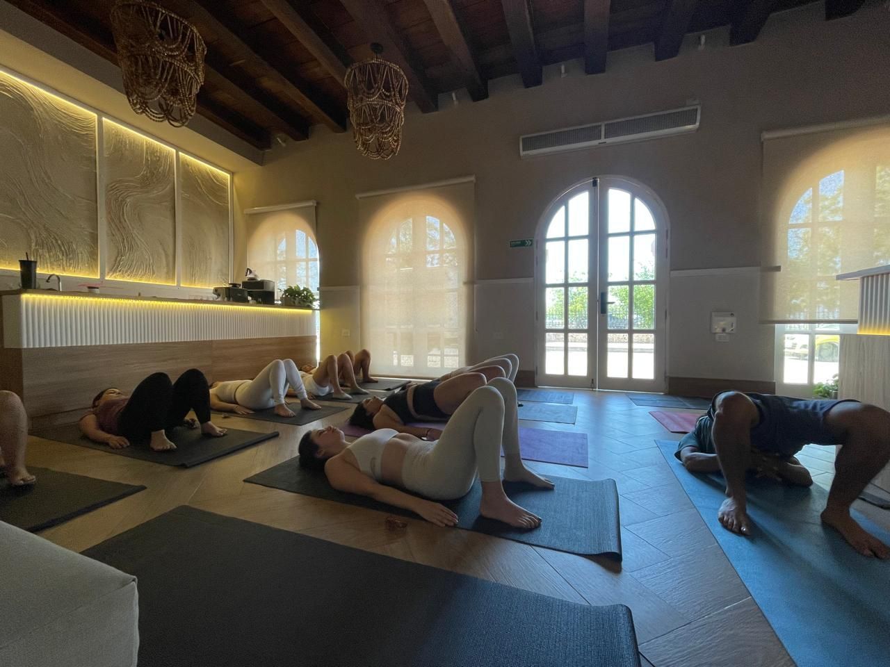 People doing yoga in a light-filled room with arched windows. They are in bridge pose on yoga mats.