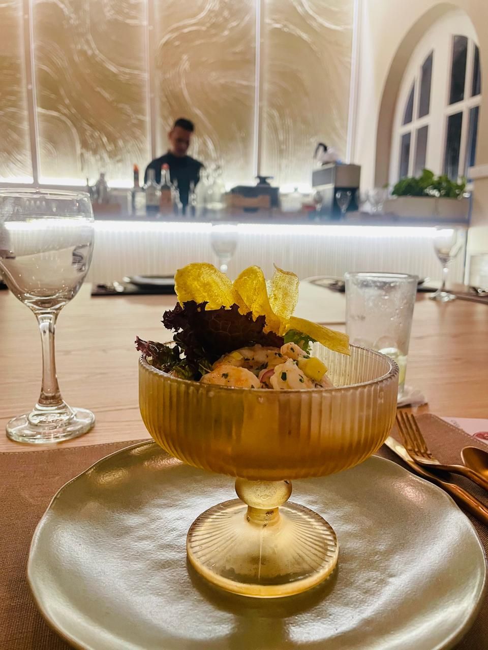 Close-up of a dessert served in a gold-tinted glass dish, on a golden plate. A restaurant bar is in the background.