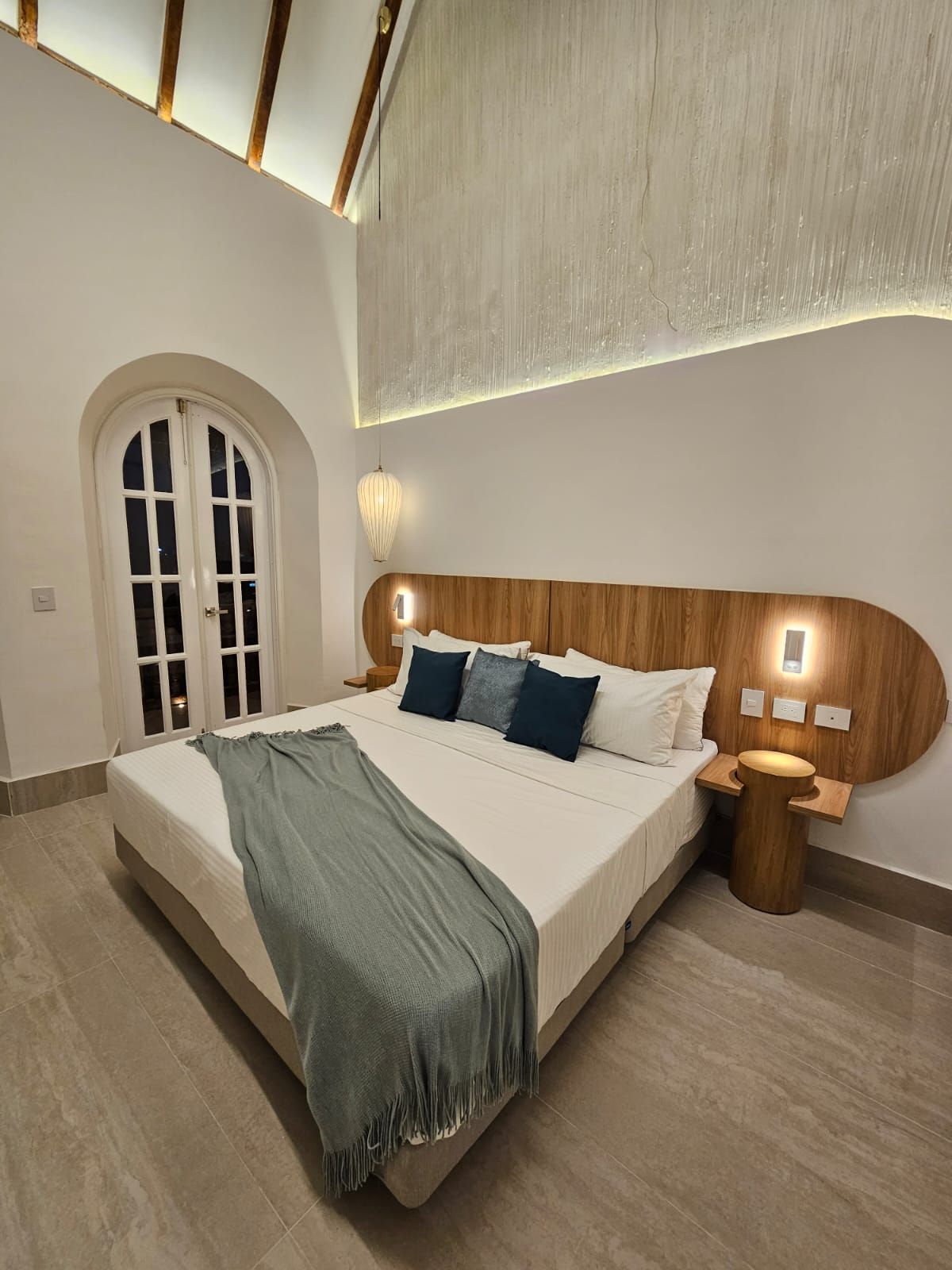 Bedroom with a large bed, wooden headboard, and side table. Arched French doors and a high ceiling are also visible.
