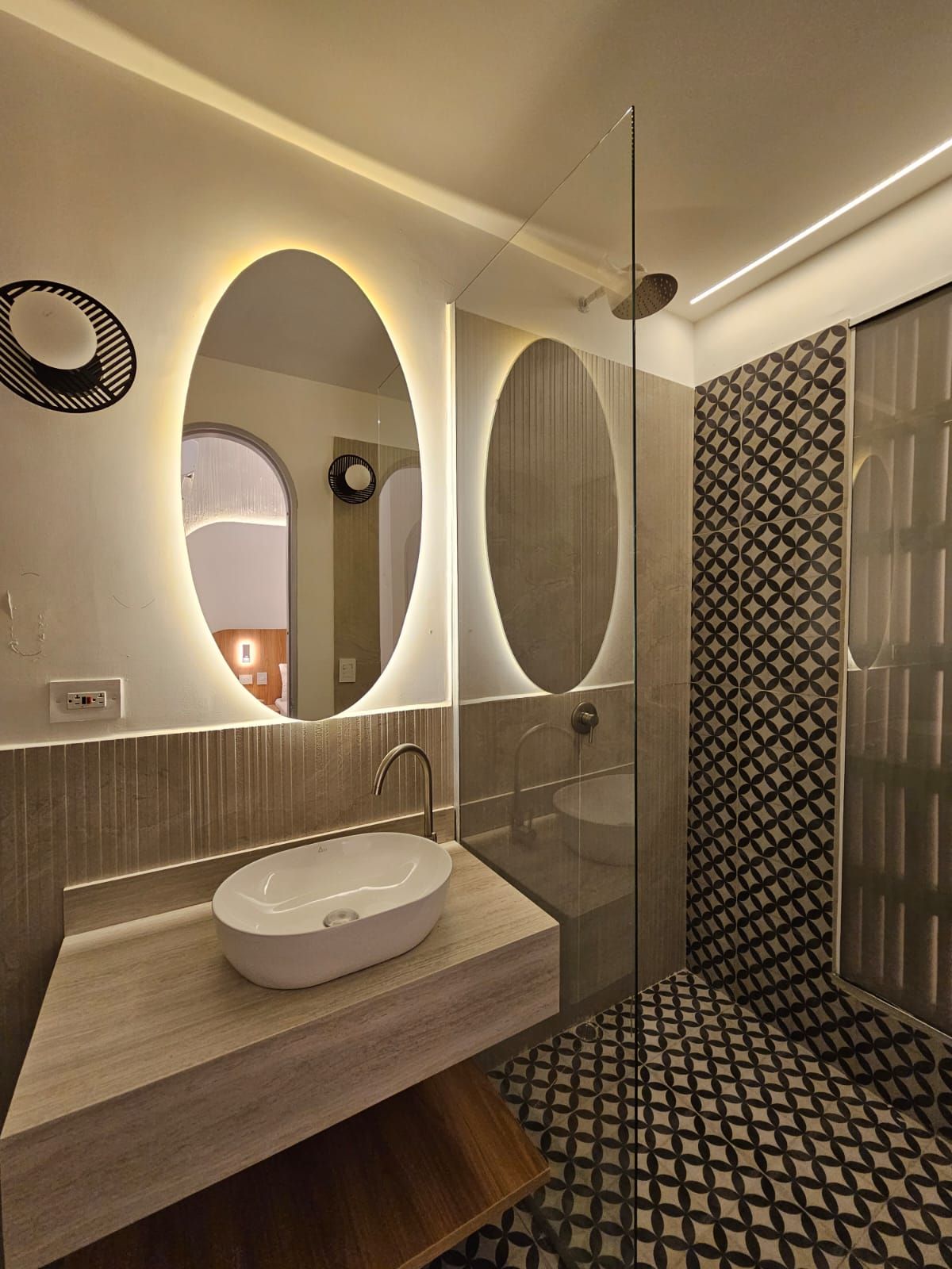 A modern bathroom with an oval-shaped mirror, a vessel sink, and a patterned tile shower. Warm lighting highlights the space.