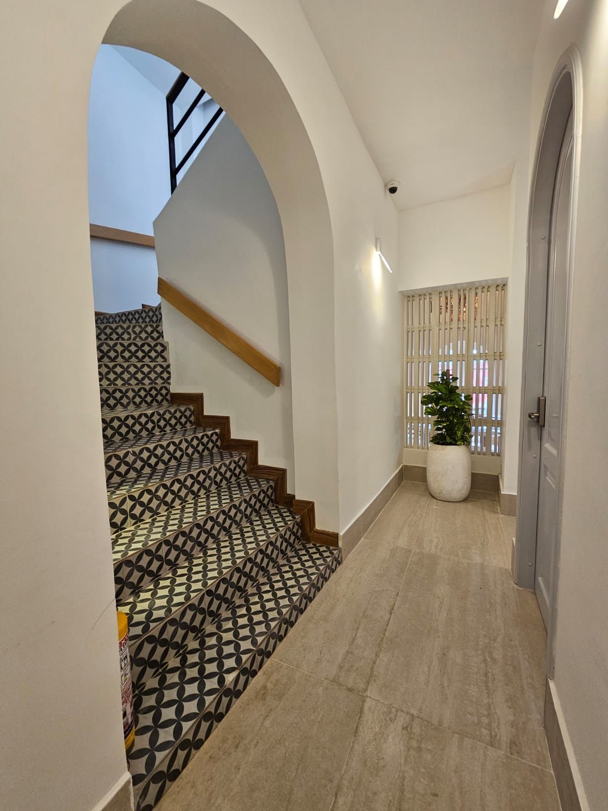 Hallway with patterned staircase on the left, wooden floor, and a doorway on the right. A potted plant sits near a window.