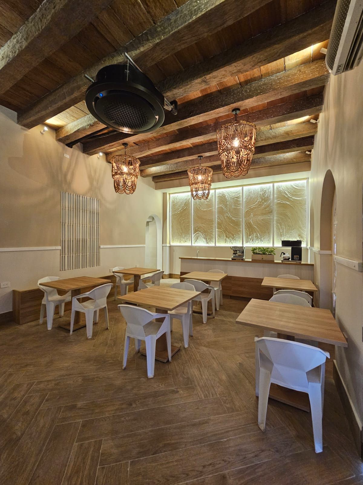 An empty restaurant interior with wooden tables and white chairs, beneath a wood-beamed ceiling and decorative hanging lights.