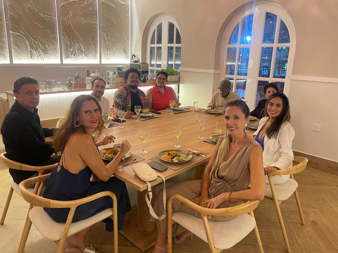 Group of people at a table in a restaurant, smiling and enjoying a meal. Warm lighting, neutral tones, and arched windows are visible.
