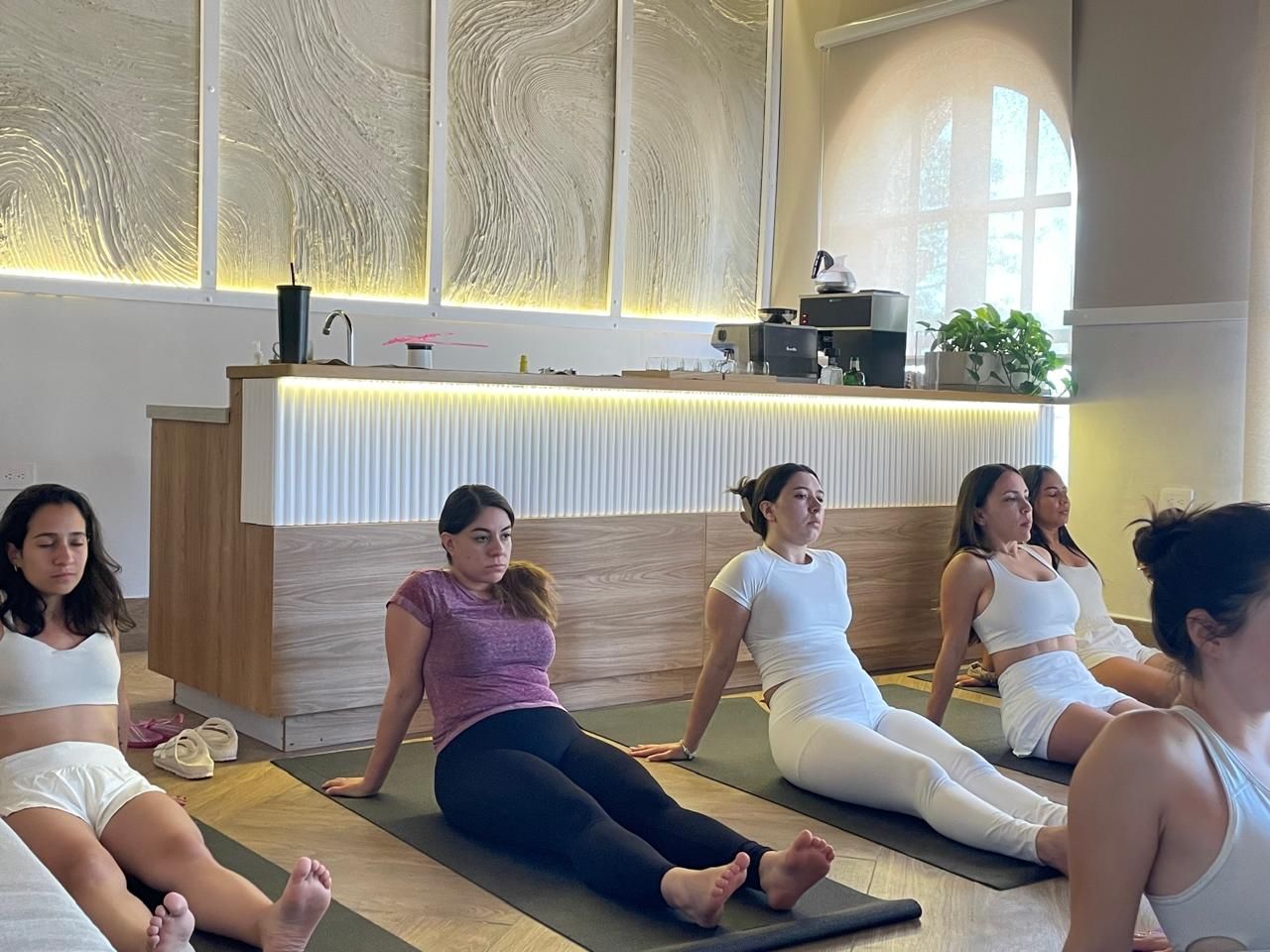 People sitting on yoga mats, stretching, in a bright room. Some wear white, others dark leggings. Light wood and white wall details are behind them.