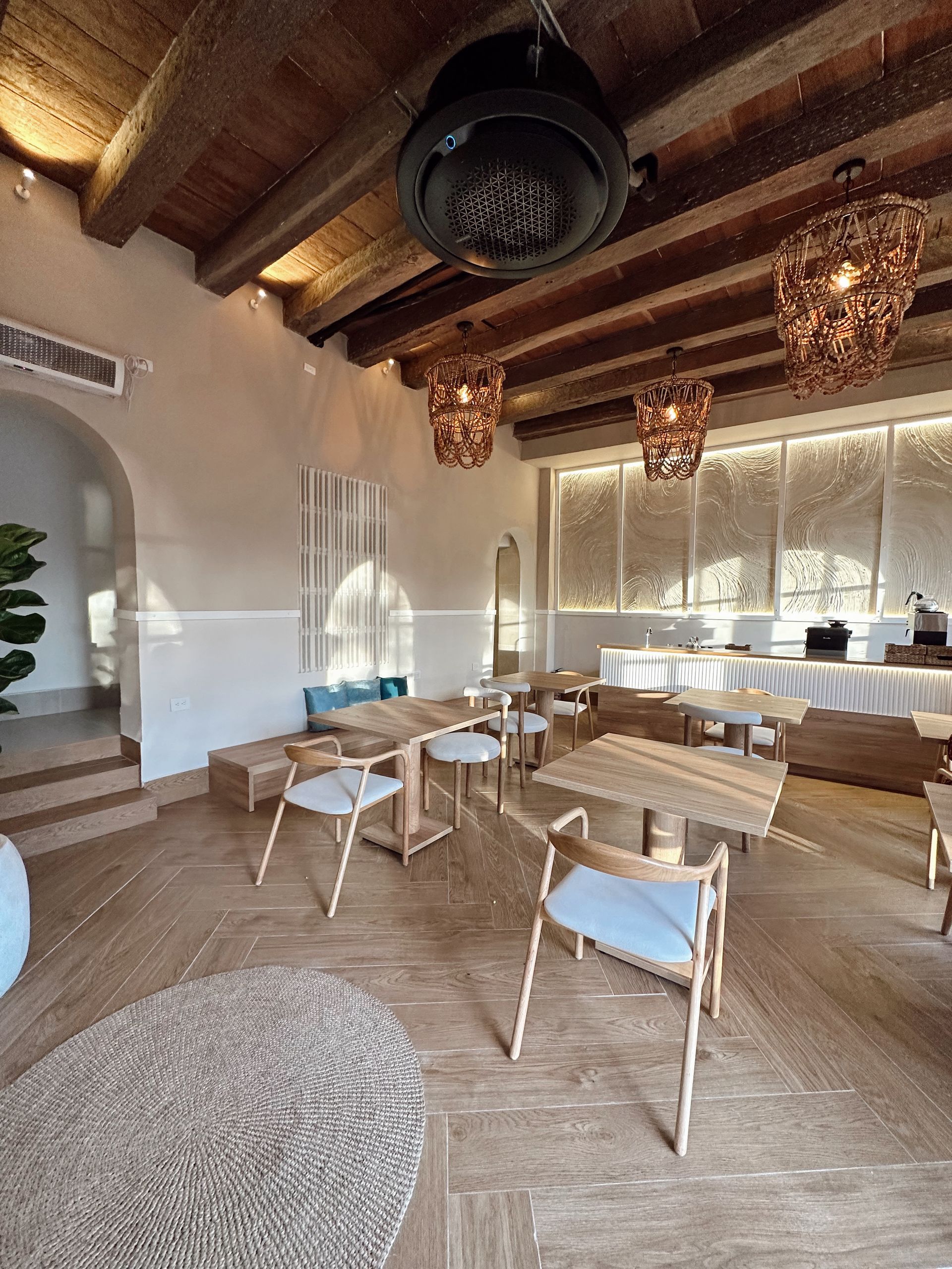 Interior of a restaurant with wooden tables and chairs. Decorative lamps hang from a wood-beam ceiling. Sunlight streams in through windows.