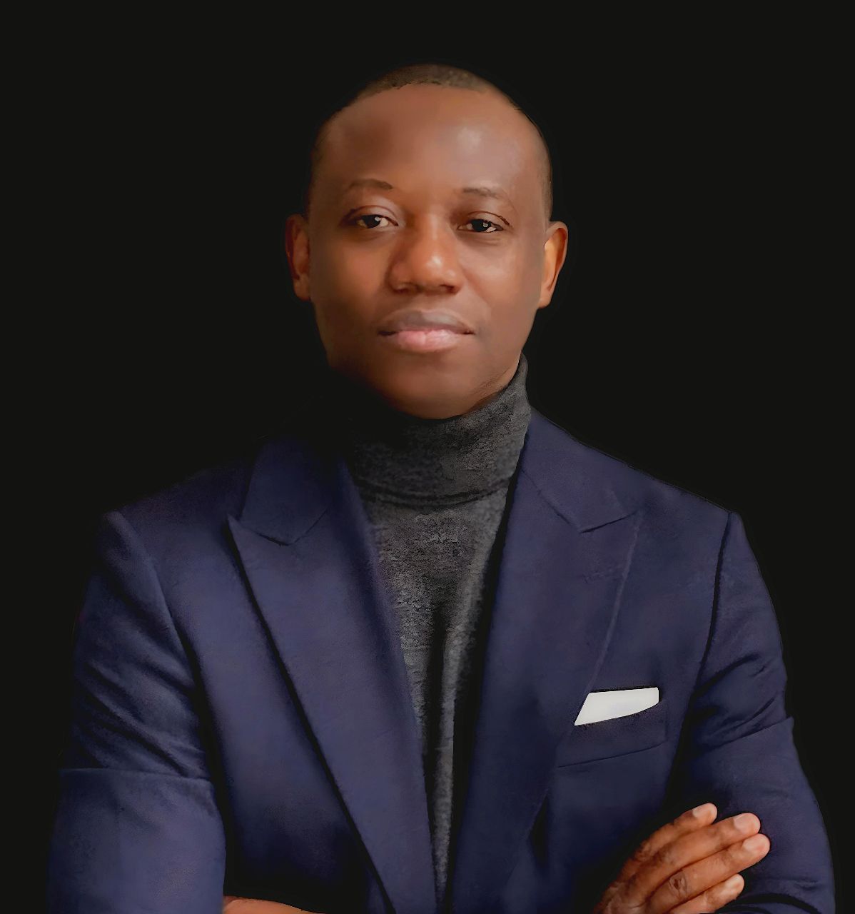 Studio portrait of a person in a navy blazer and dark turtleneck, arms crossed against a black background