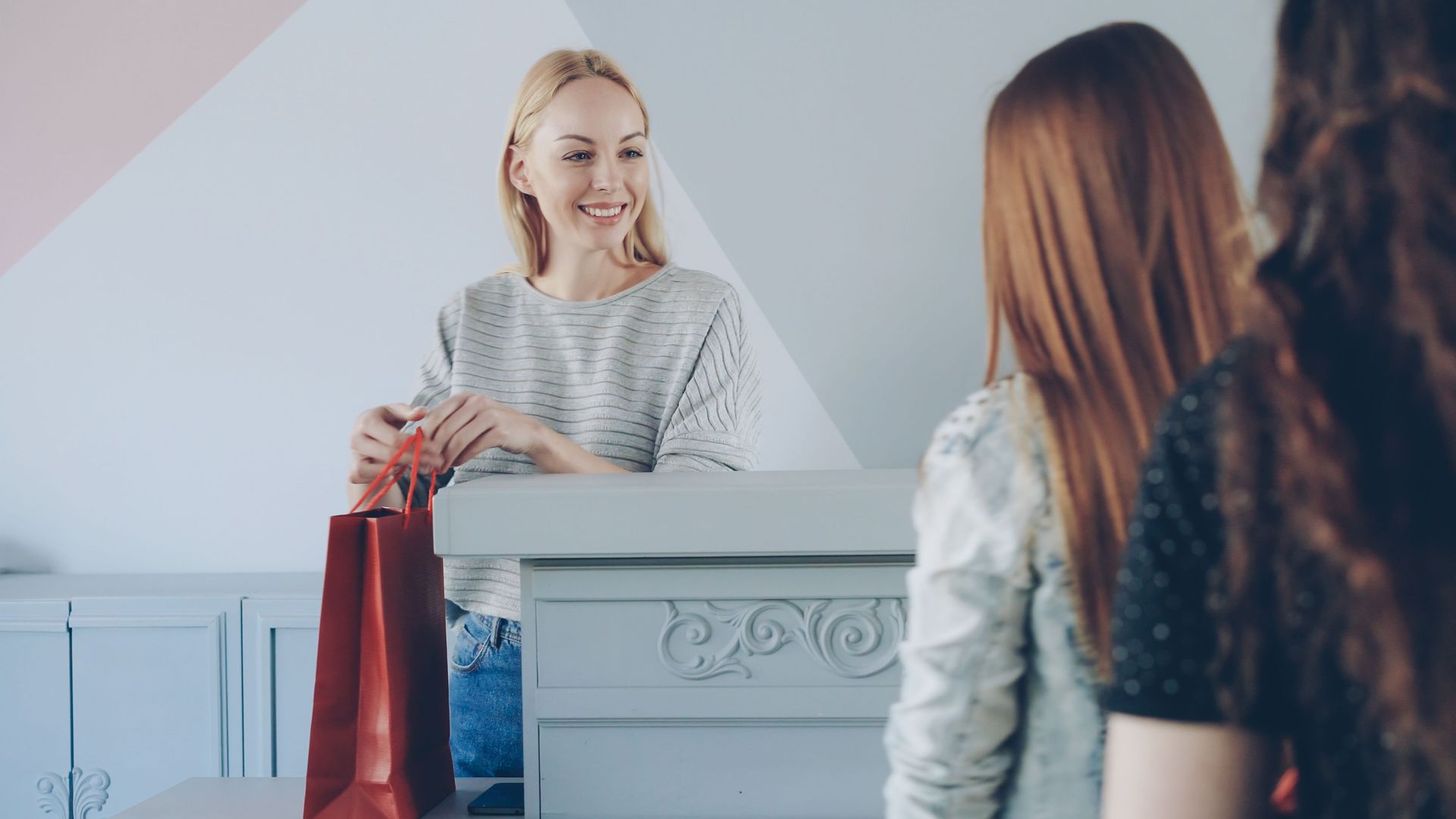 Blonde shop clerk smiling at a counter, handing a red shopping bag to a customer in a bright store