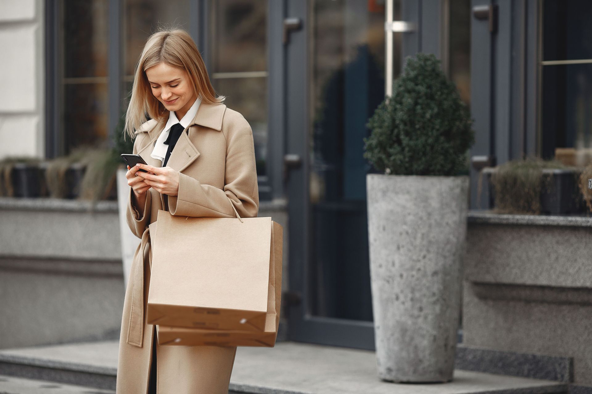Woman in a beige coat checking her phone beside a shopping bag outside a storefront.
