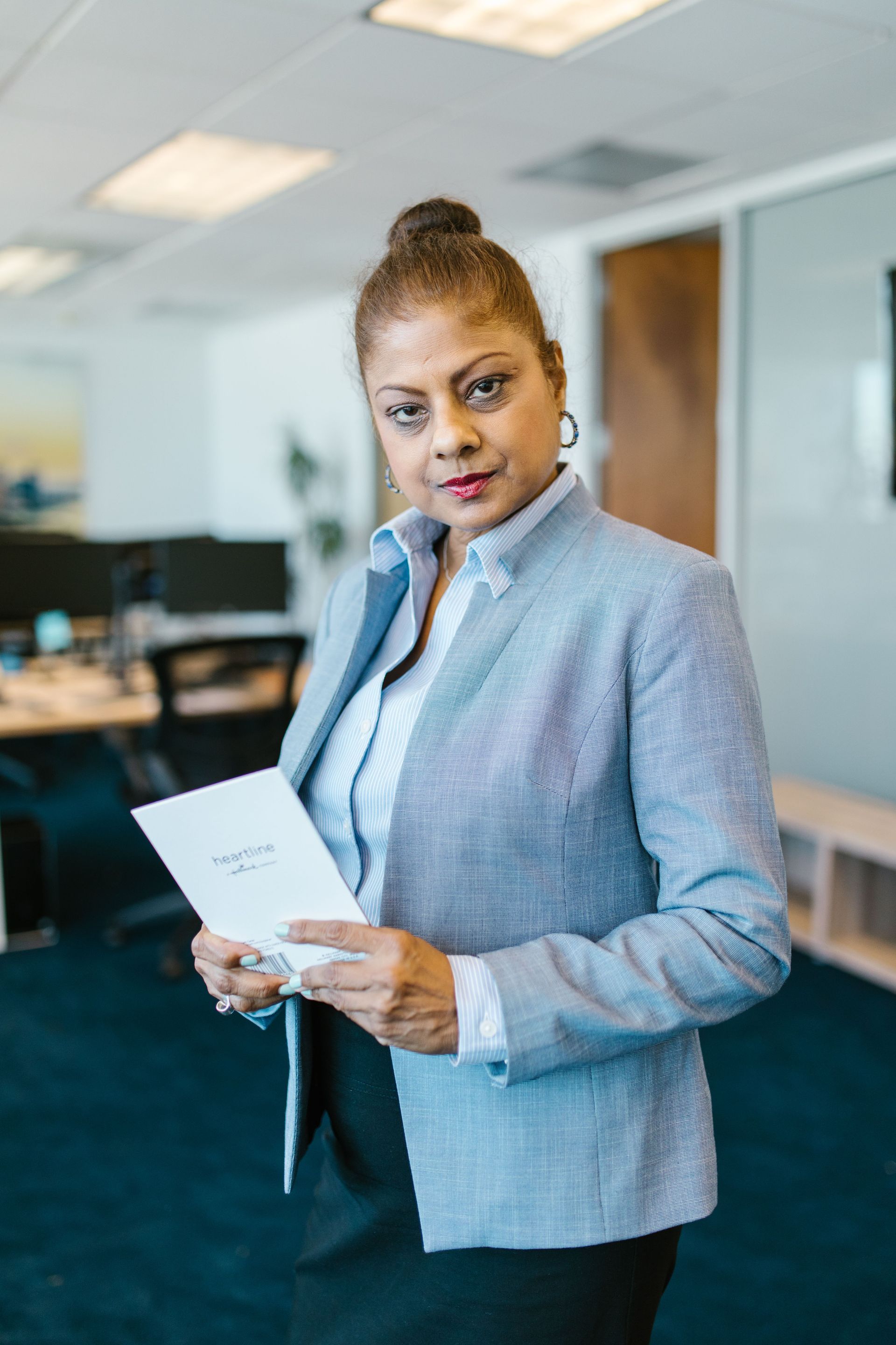 Woman in a light gray blazer holds a paper in a modern office hallway, looking at the camera.