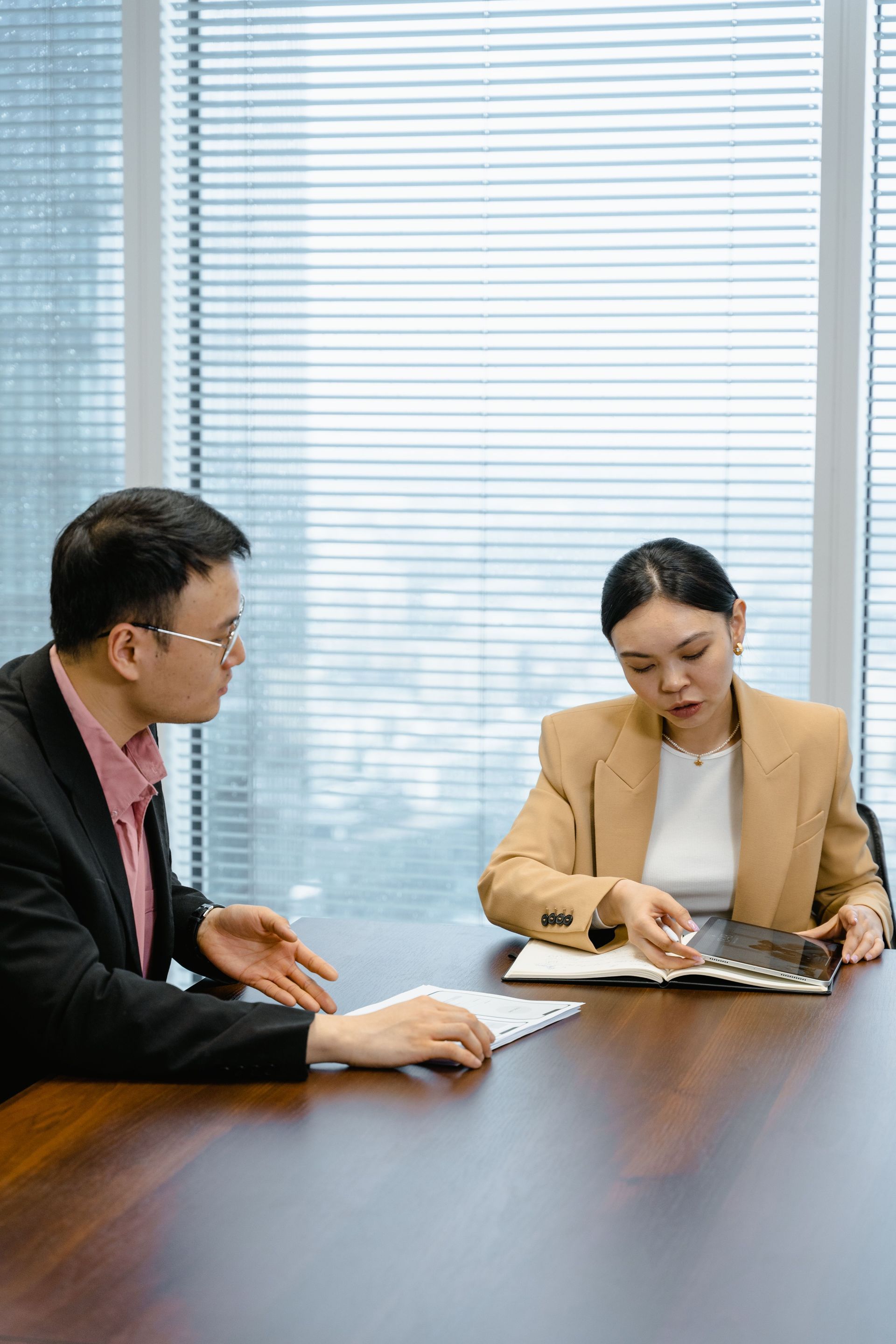 Two colleagues reviewing documents at a conference table in an office, with blinds behind them.
