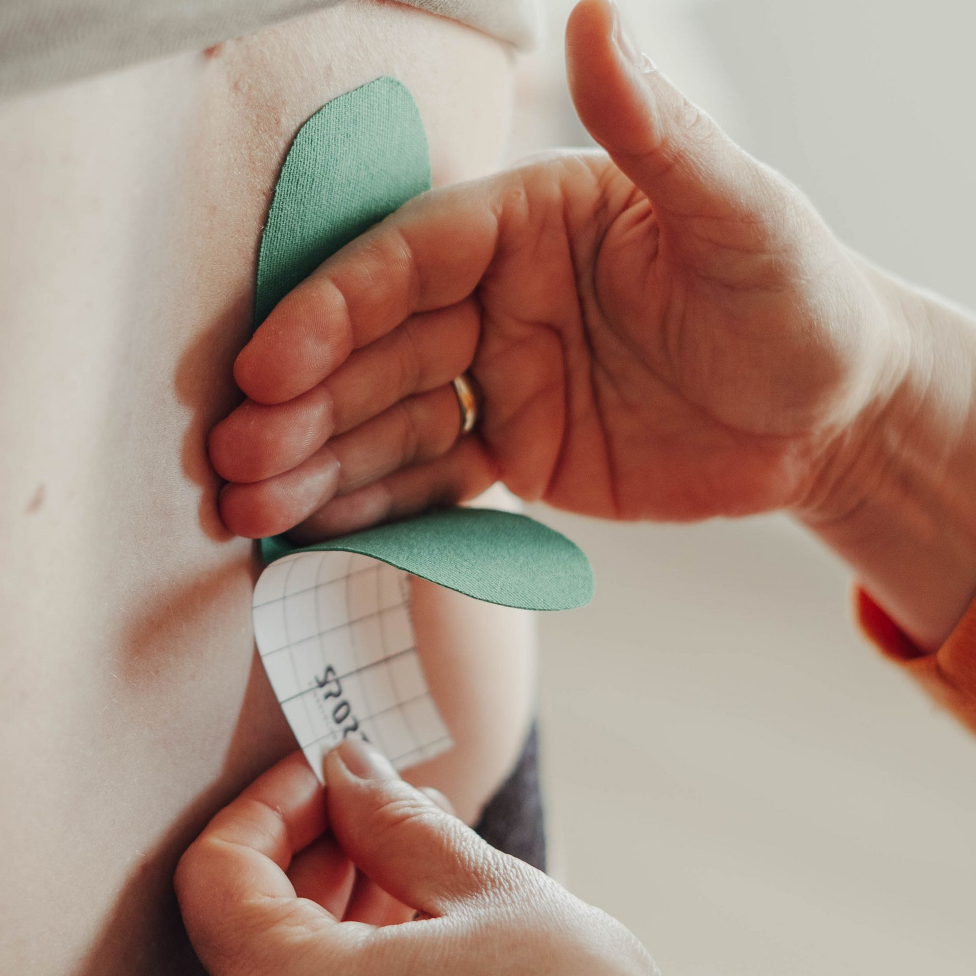 Eine Person klebt grünes Kinesiologie-Tape auf den Rücken. Die Hand zieht die Schutzfolie des Tapes ab.