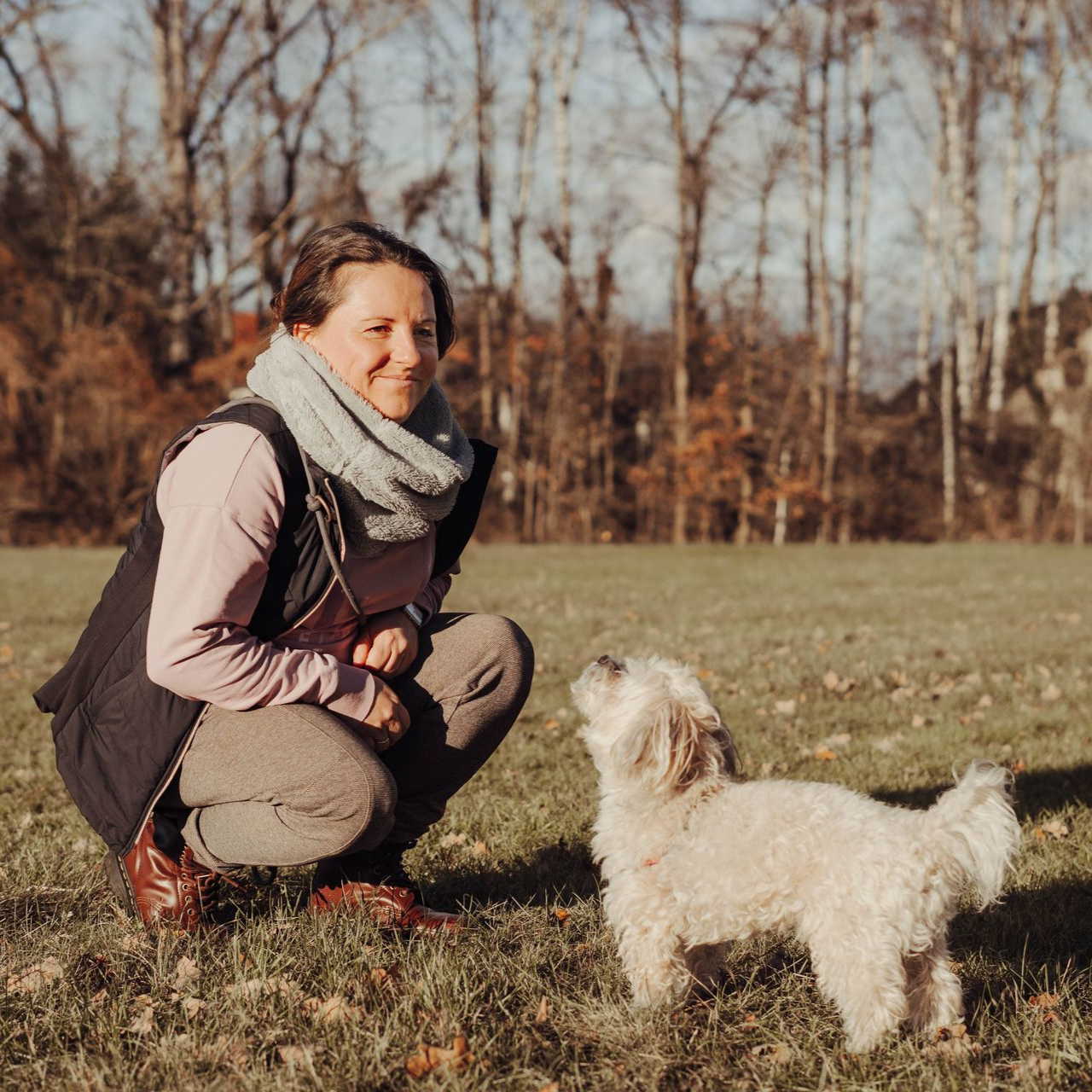 Anja Täubert, herbstlich angezogen und hockend, betrachtet einen kleinen, flauschigen Hund auf einer Wiese im Herbst.