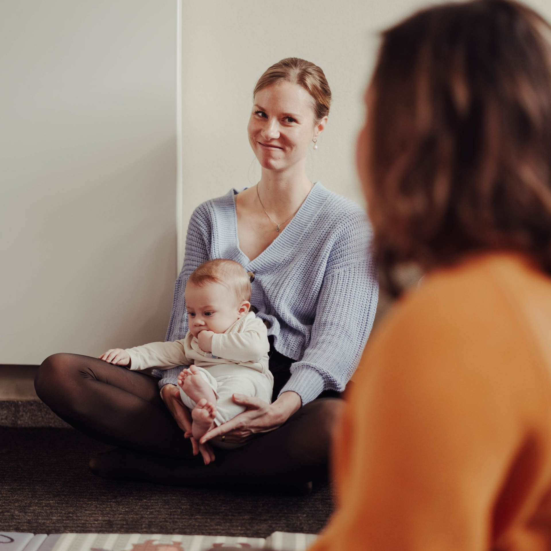 Frau sitzt mit Baby in einem Zimmer und blickt lächelnd zu Anja Täubert - Hebamme und Stillberaterin.