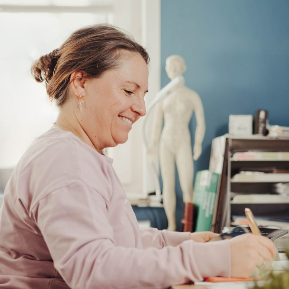 Frau in rosa Pullover lächelt beim Schreiben an einem Schreibtisch; im Hintergrund ist eine blaue Wand zu sehen.