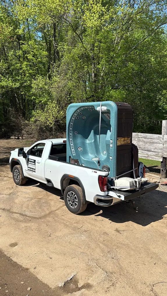 White pickup truck carrying a blue hot tub, parked on a dirt road near trees.