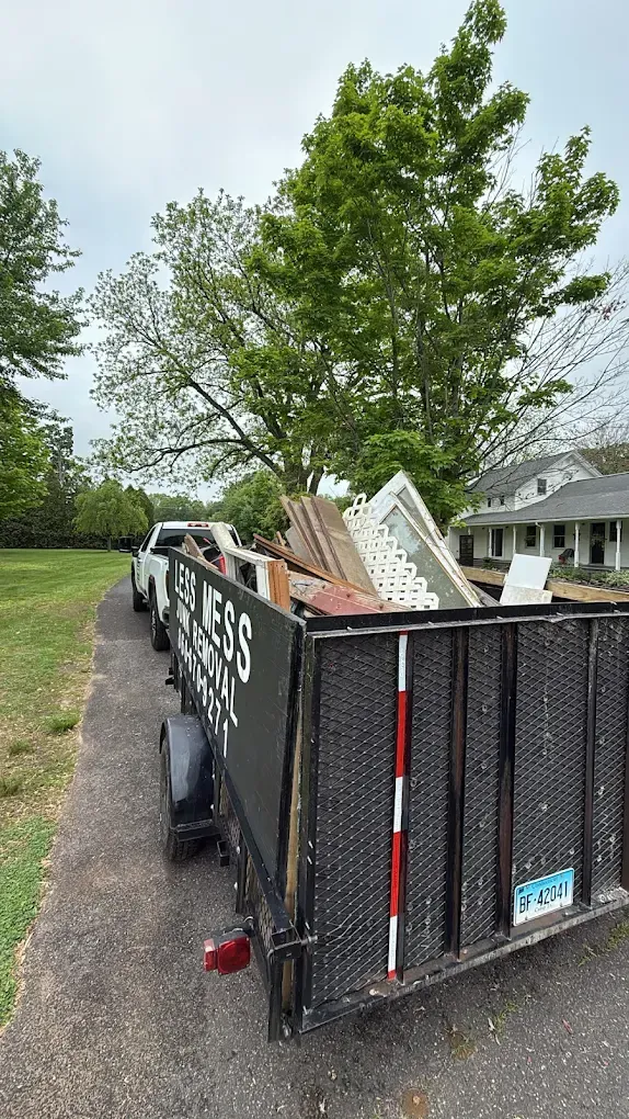 A trailer loaded with debris is hitched to a white pickup truck on a driveway next to a house.