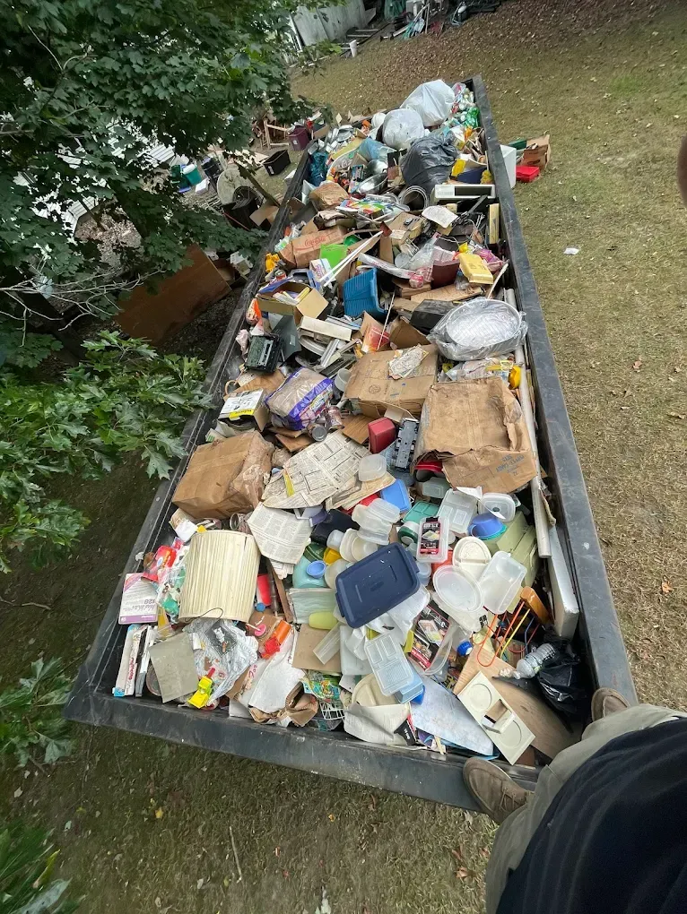 A rectangular dumpster overflowing with trash: cardboard, plastic, paper, and food scraps. Outdoors, on grass.