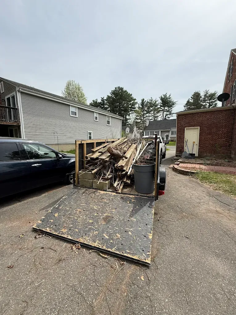 Trailer loaded with wood debris in a driveway, next to a black car and buildings.